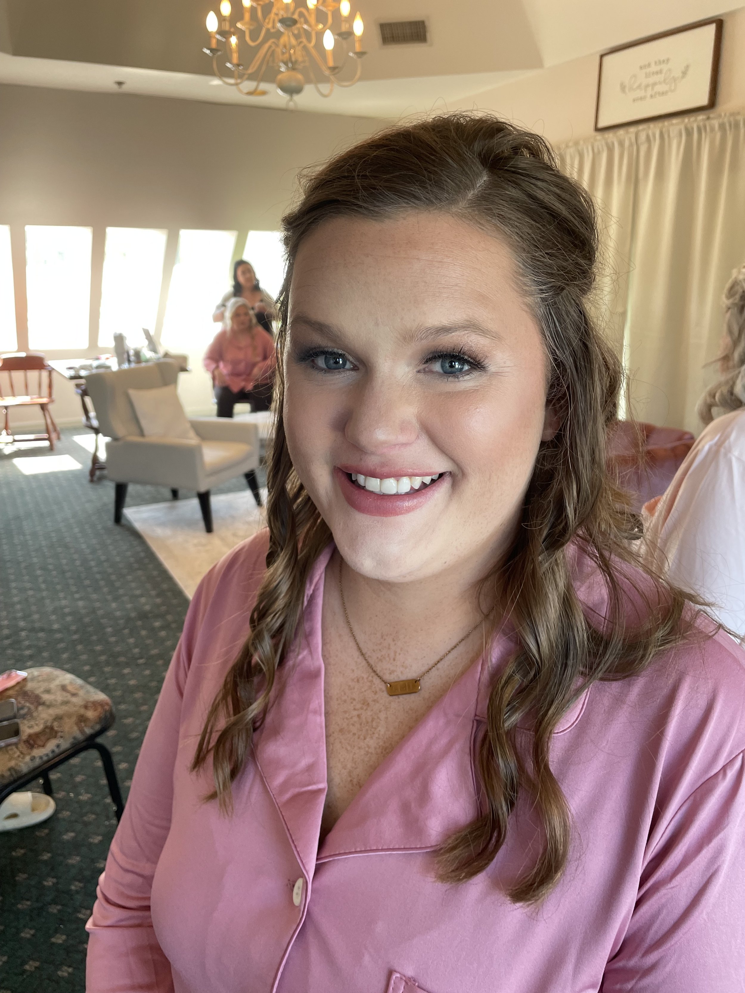 A smiling woman with curly brown hair in a pink silky top, wearing a gold necklace, in a cozy room with a chandelier, armchair, and women in the background.