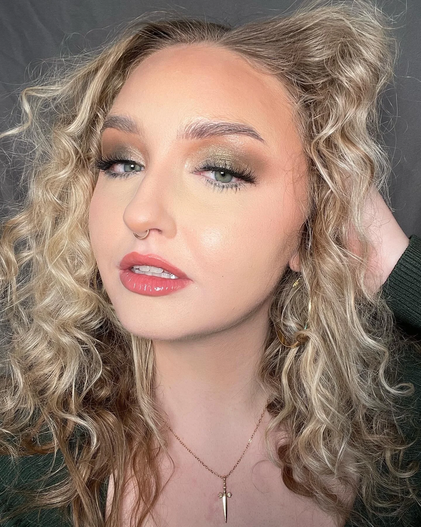 A woman with long, curly blonde hair and fair skin, wearing makeup with green eyeshadow, black eyeliner, and pink lipstick. She has a septum piercing, gold hoop earrings, and a necklace with a dagger pendant. She is posing against a dark background, 