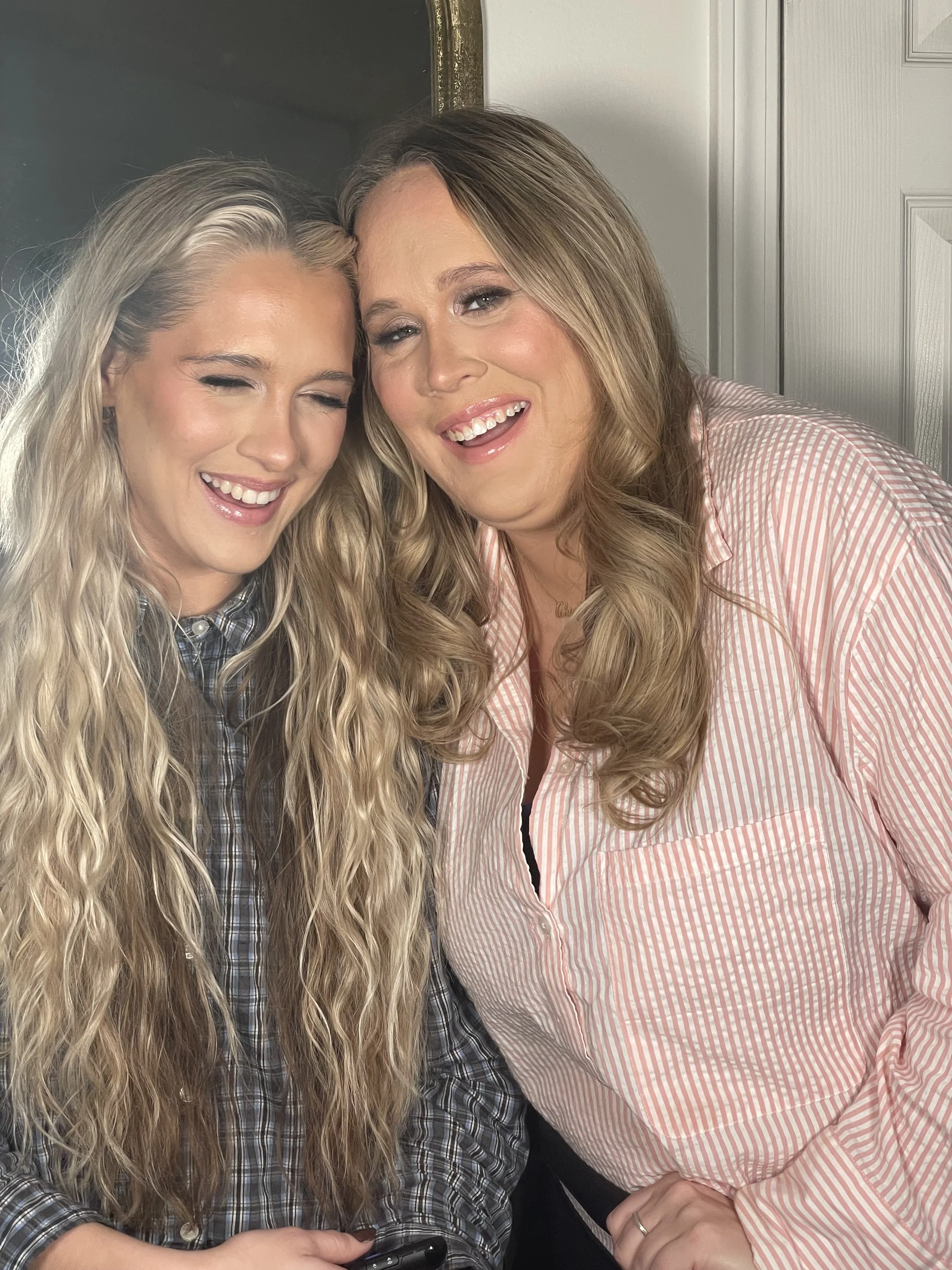 Two women smiling, close together, indoors, with brick wall and door in background.