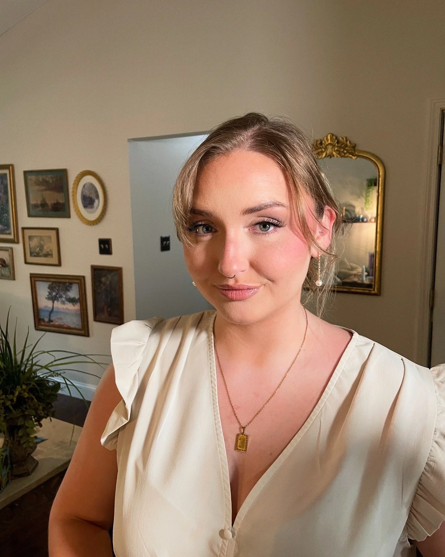 A woman with light brown hair and blue eyes smiling slightly, wearing a beige top with puff sleeves, gold jewelry including a pendant necklace and dangling earrings, in a room with framed pictures on the wall and a gold-framed mirror.