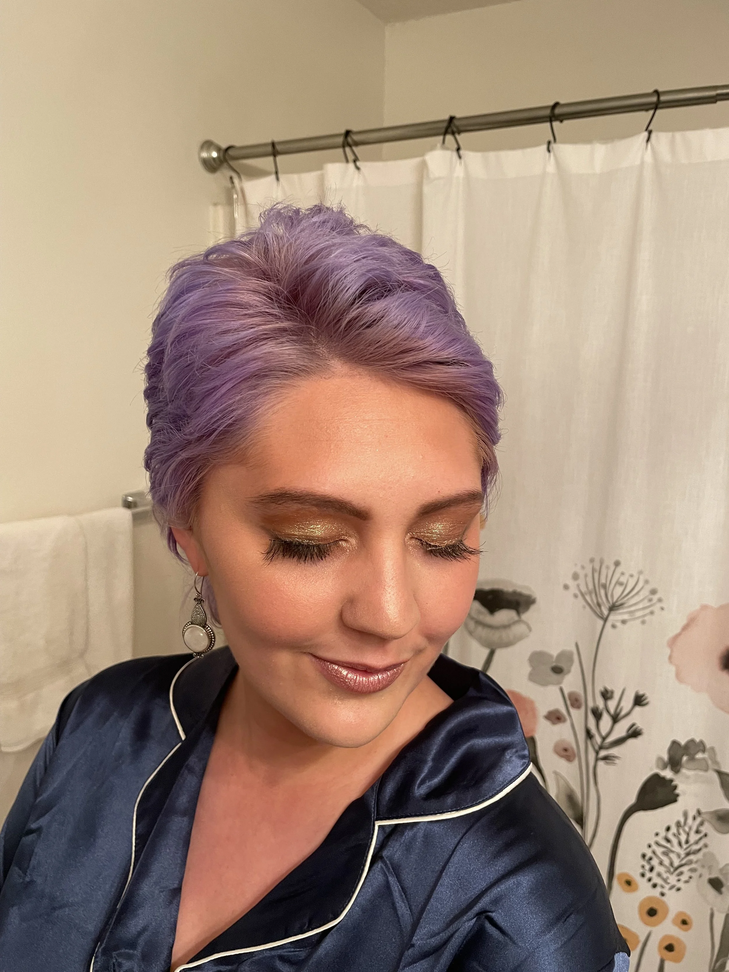 Woman with short purple hair smiling with makeup in a bathroom, wearing a navy satin pajama top and dangle earrings, with floral wallpaper and a shower curtain in the background.