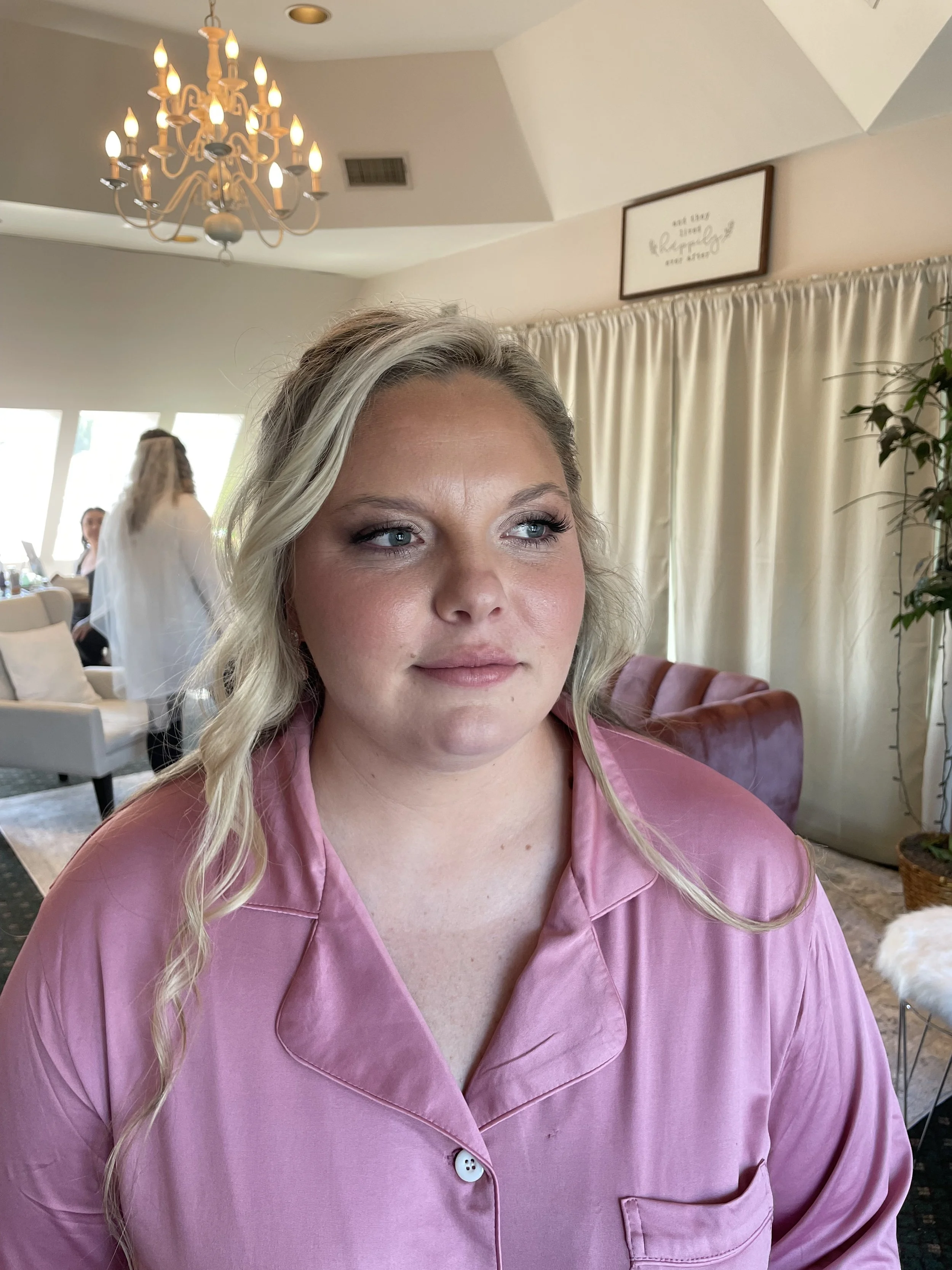 A woman with blonde hair styled in loose waves, wearing pink satin pajamas, sitting indoors near a cream-colored curtain. In the background, there is a chandelier, and some people can be seen in a bright sitting area.