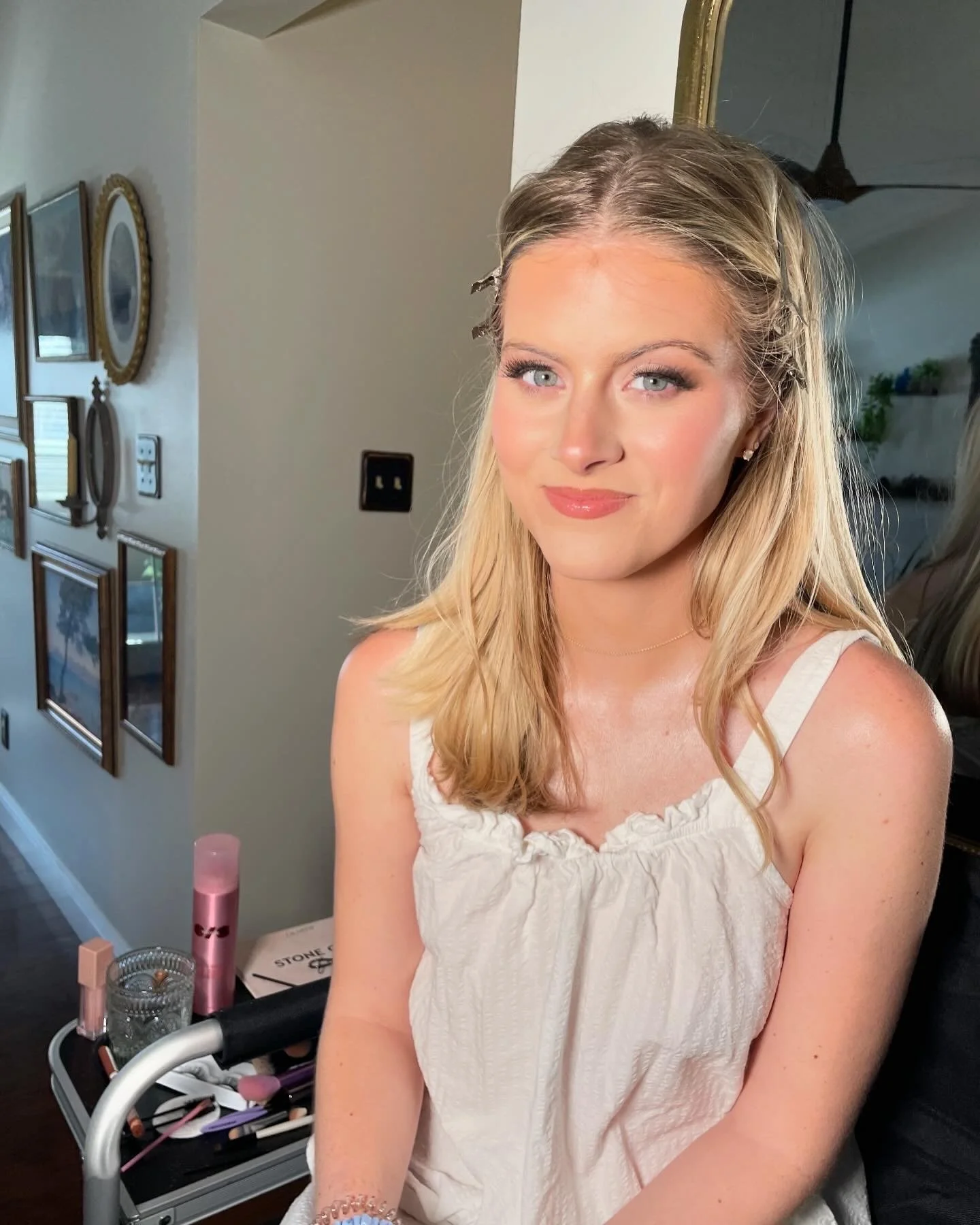 A young woman with blonde hair and blue eyes sitting indoors, wearing a white sleeveless top, with a calm and confident expression.