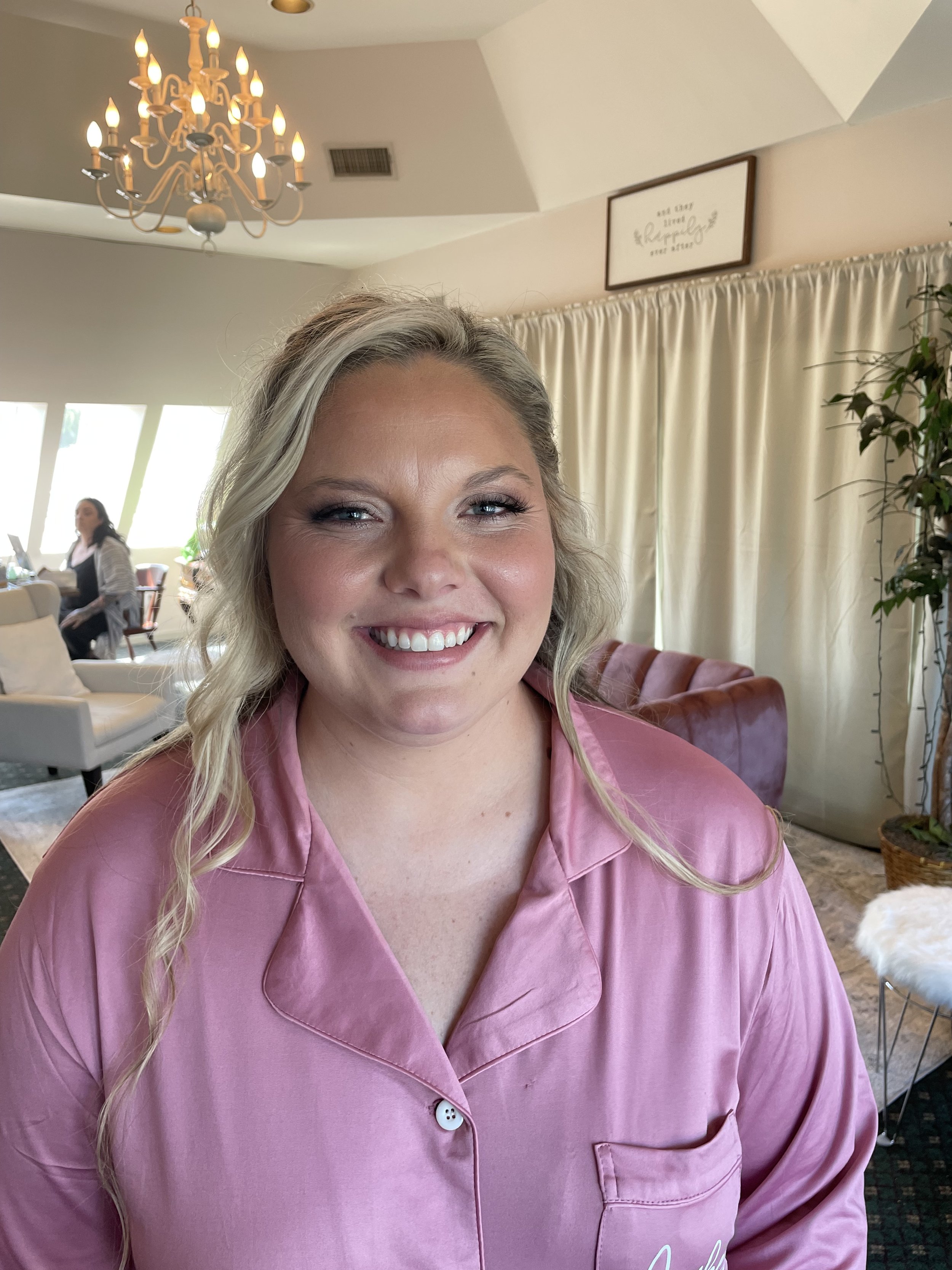 A woman with blonde hair and a pink satin pajama top smiling in a well-lit living room with a chandelier, curtains, and other people in the background.