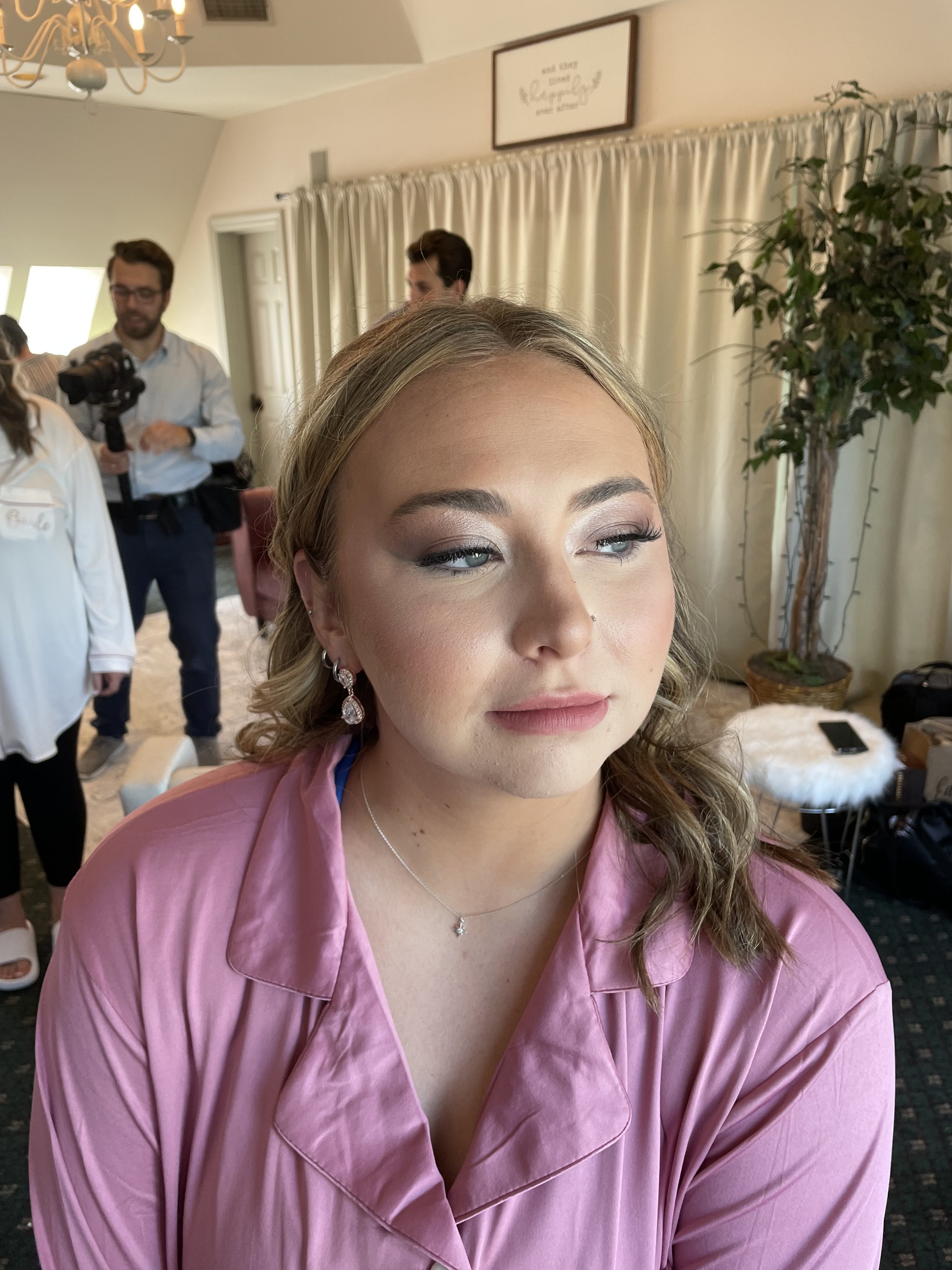 A young woman with blonde hair and makeup, wearing pink satin pajamas and jewelry, sitting indoors with a neutral expression.