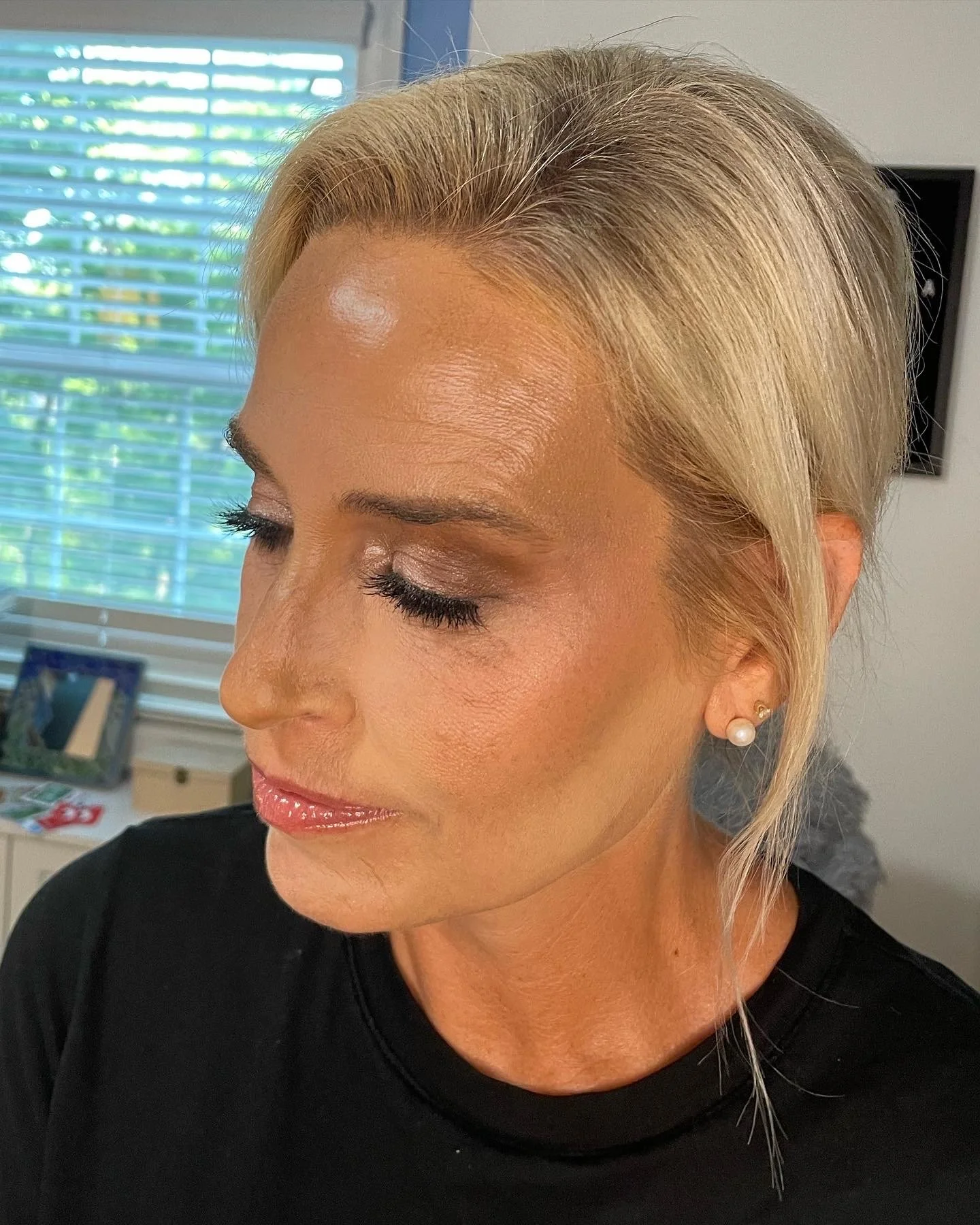Close-up of a woman's face with blonde hair, makeup, and pearl earrings, indoors with blinds and a window in the background.