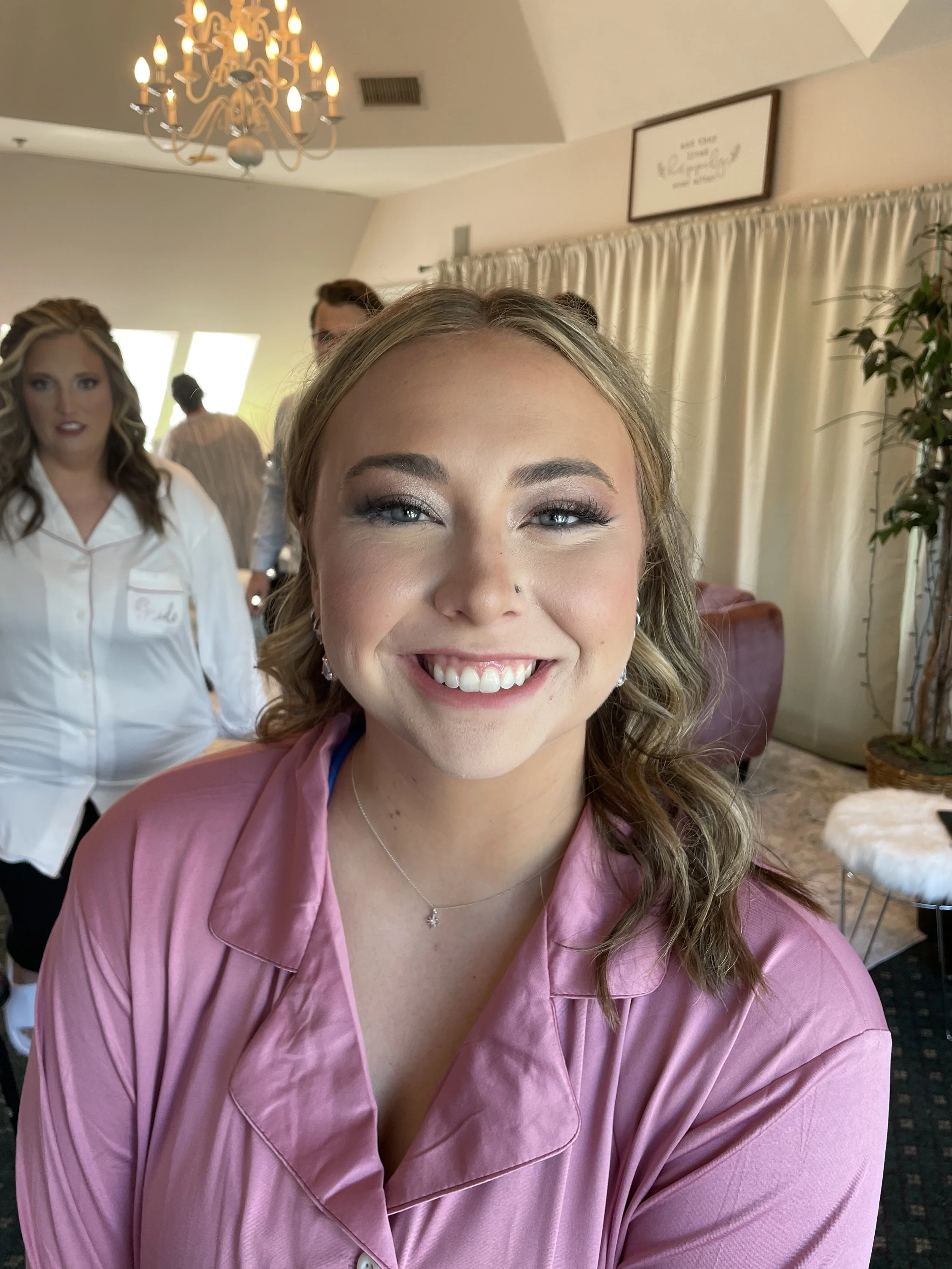 A smiling woman with light brown, wavy hair, wearing a pink satin pajama top and jewelry, standing indoors with a chandelier, curtains, and other people in the background.