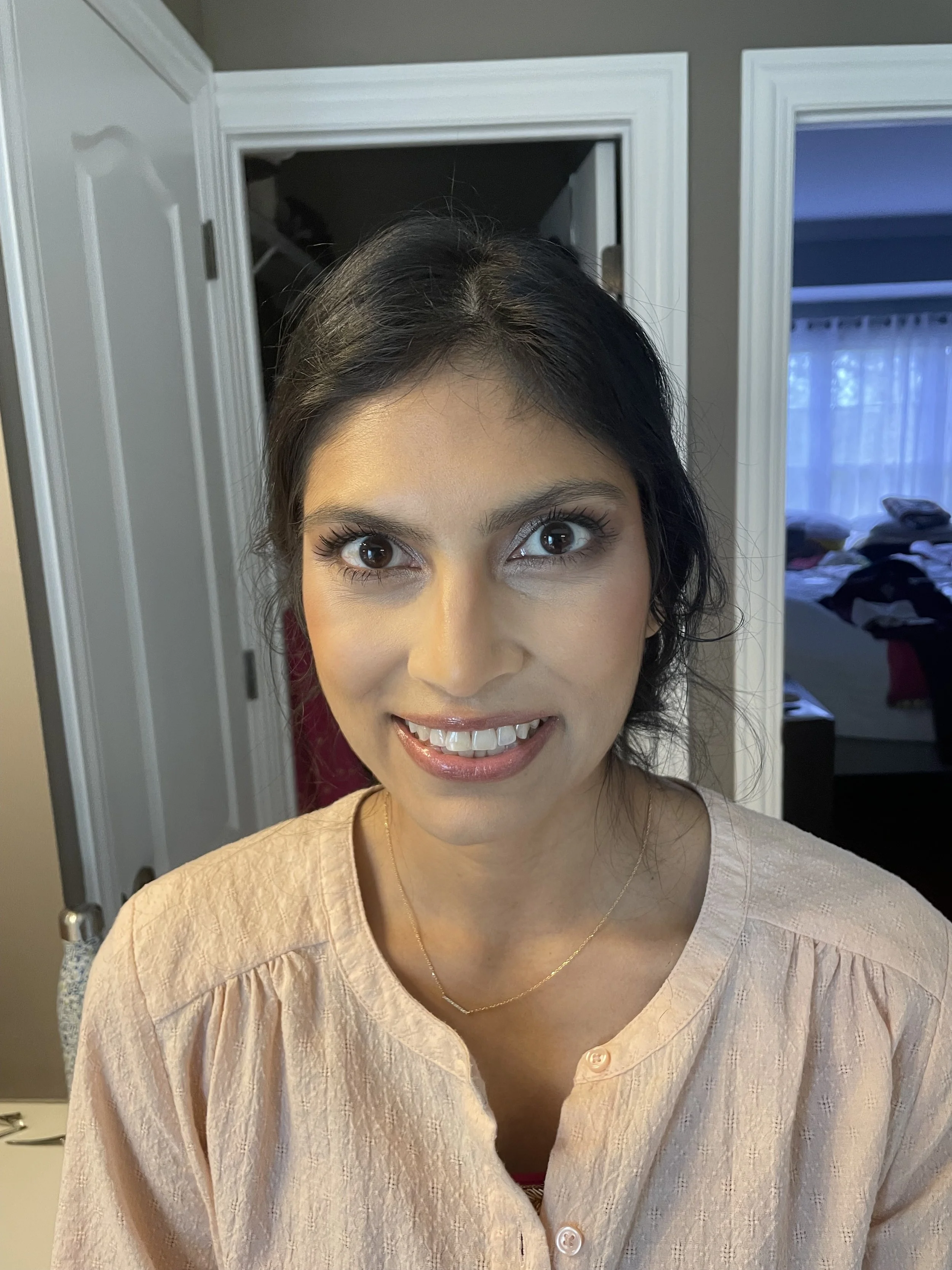 Close-up of a woman with dark hair, wearing makeup and a light-colored blouse, smiling at the camera indoors with a bedroom and closet in the background.