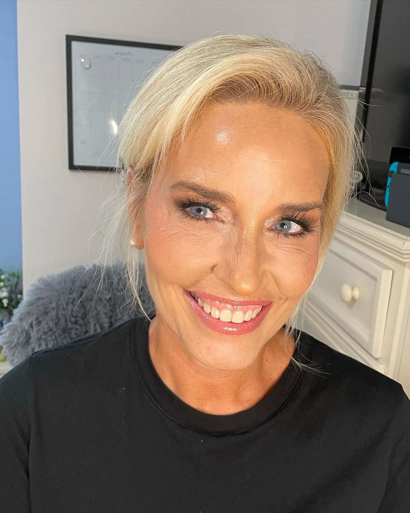 A woman taking a selfie indoors, smiling with bright teeth and wearing makeup, blonde hair, and a black top.