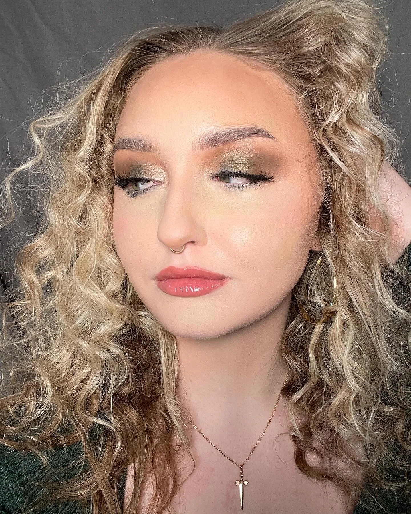 Close-up portrait of a woman with curly blonde hair, wearing gold eyeshadow, winged eyeliner, glossy coral lipstick, hoop earrings, a septum piercing, and a necklace with a dagger pendant.