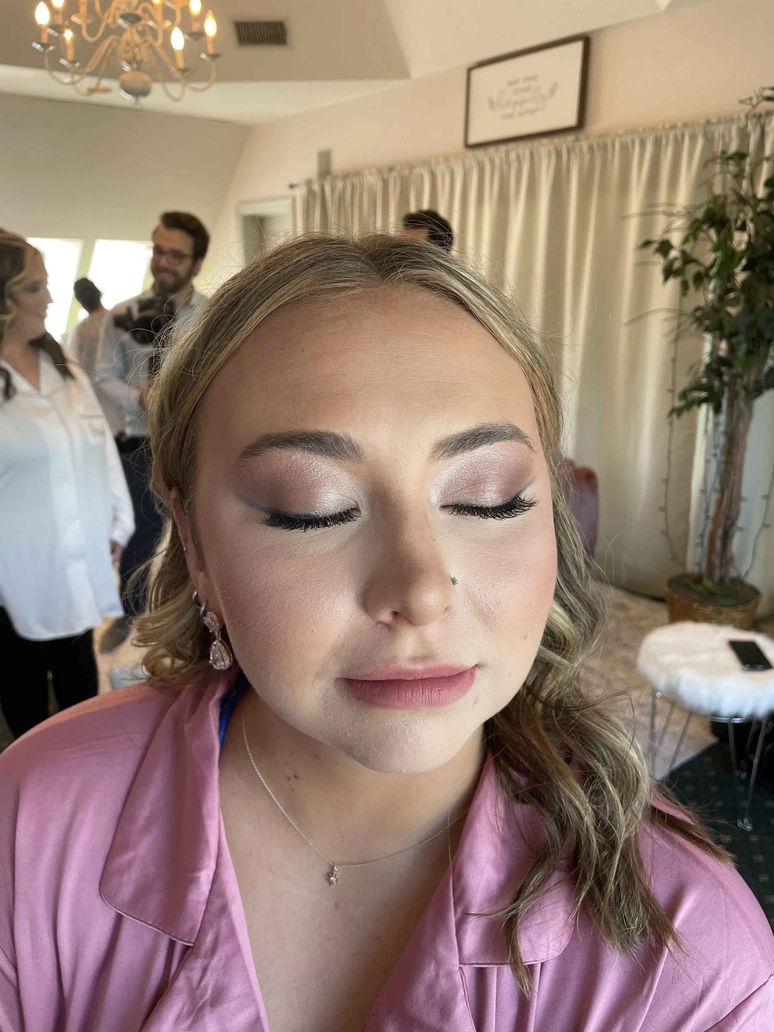 A young woman with closed eyes and makeup, wearing a pink satin robe, getting ready in a room with a chandelier, curtain, and people in the background.