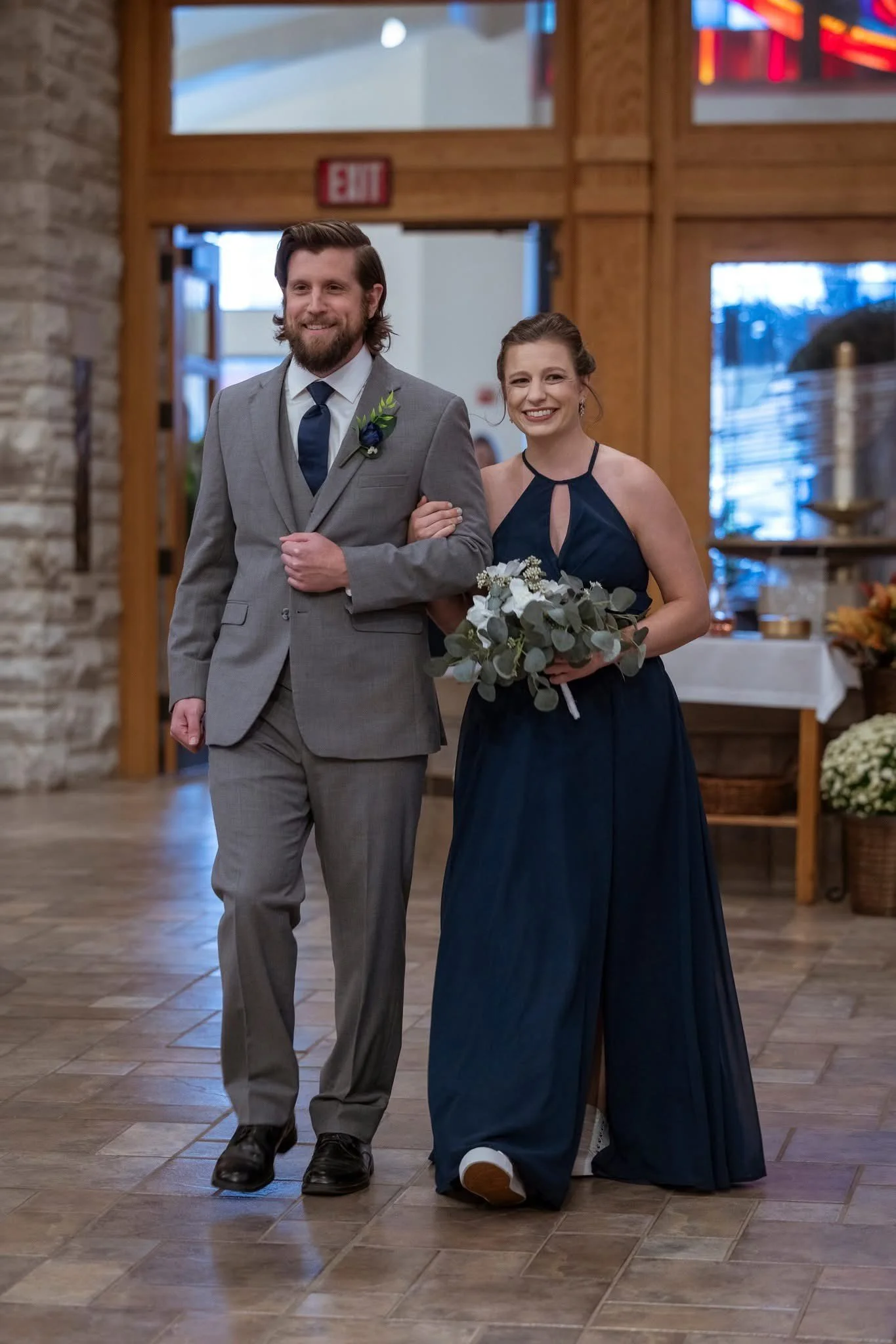 A smiling woman in a navy blue dress holding a bouquet, walking arm-in-arm with a bearded man in a gray suit, in a rustic indoor setting with wooden accents and flowers.