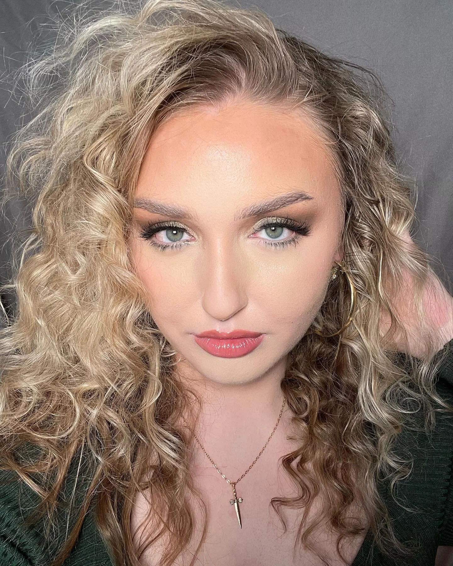 Close-up of a woman with curly blonde hair, blue eyes, wearing makeup, a black top, and a gold necklace with a dagger pendant, against a gray background.