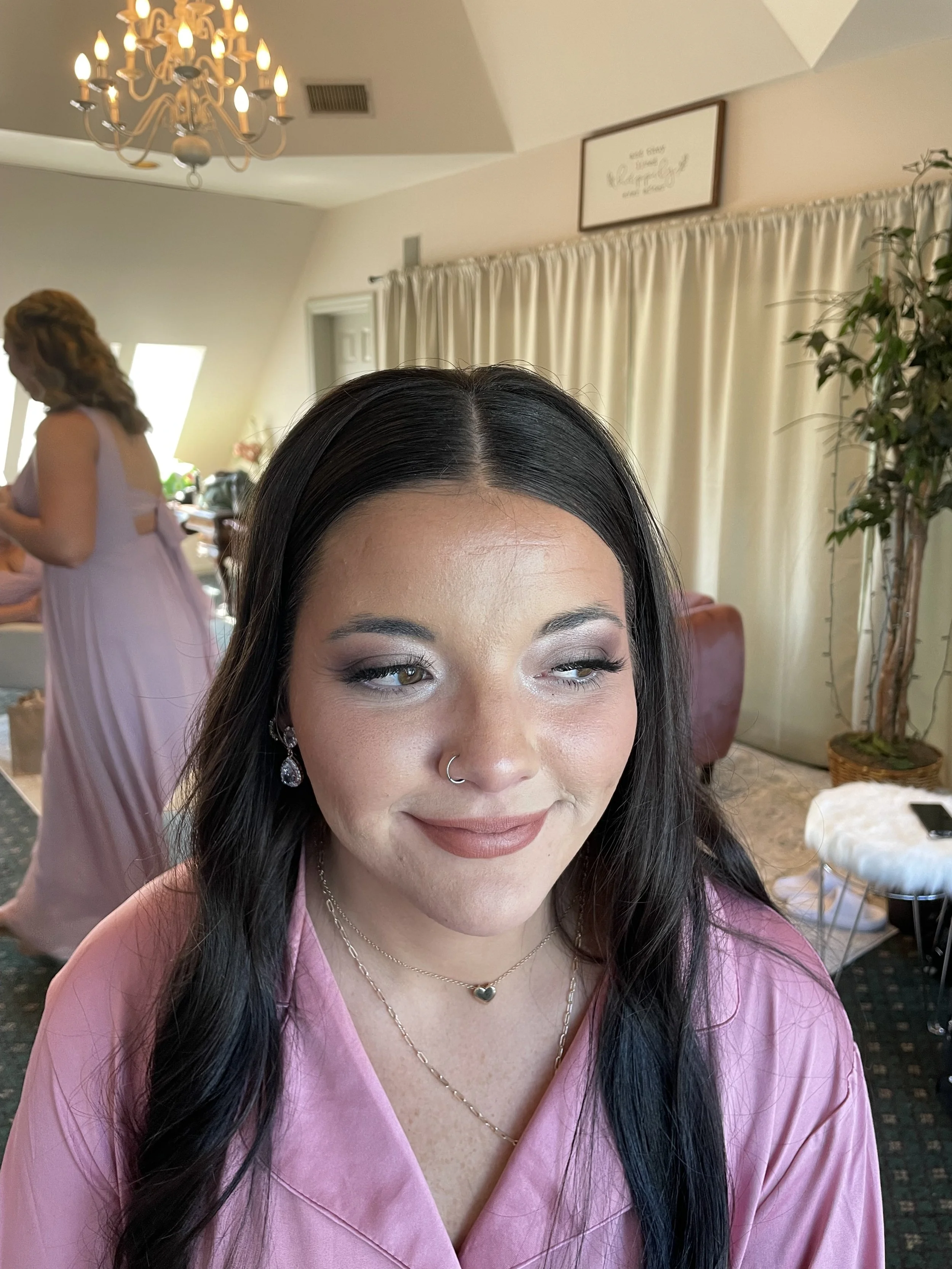 Close-up of a woman with dark hair, makeup, and a nose ring, wearing pink attire and jewelry, smiling slightly with one eye partially closed, in a room with beige curtains, a chandelier, and another woman in the background.