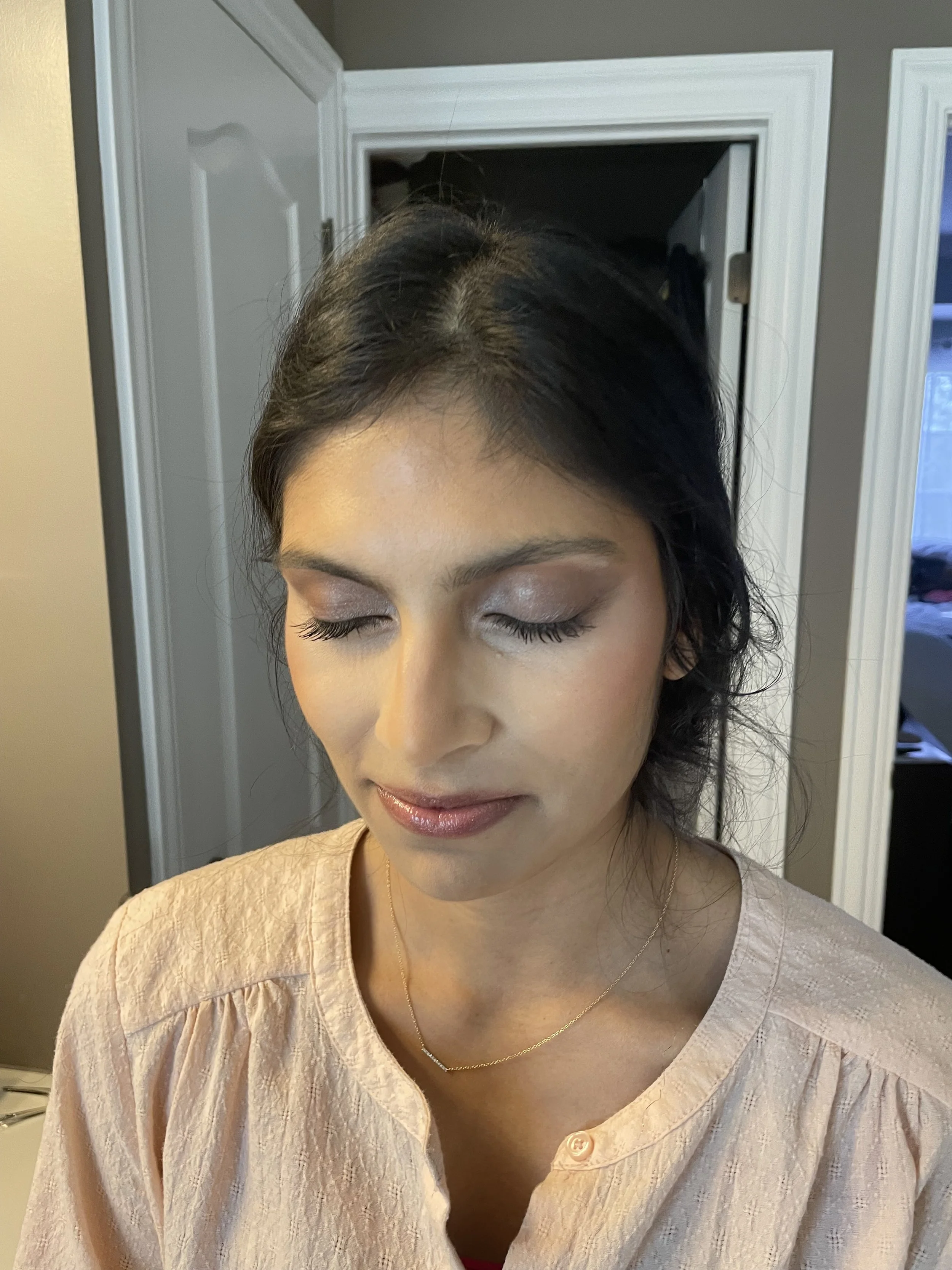 A woman with dark hair and makeup, wearing a light-colored top and a small necklace, with her eyes closed, standing in a room with an open closet behind her.