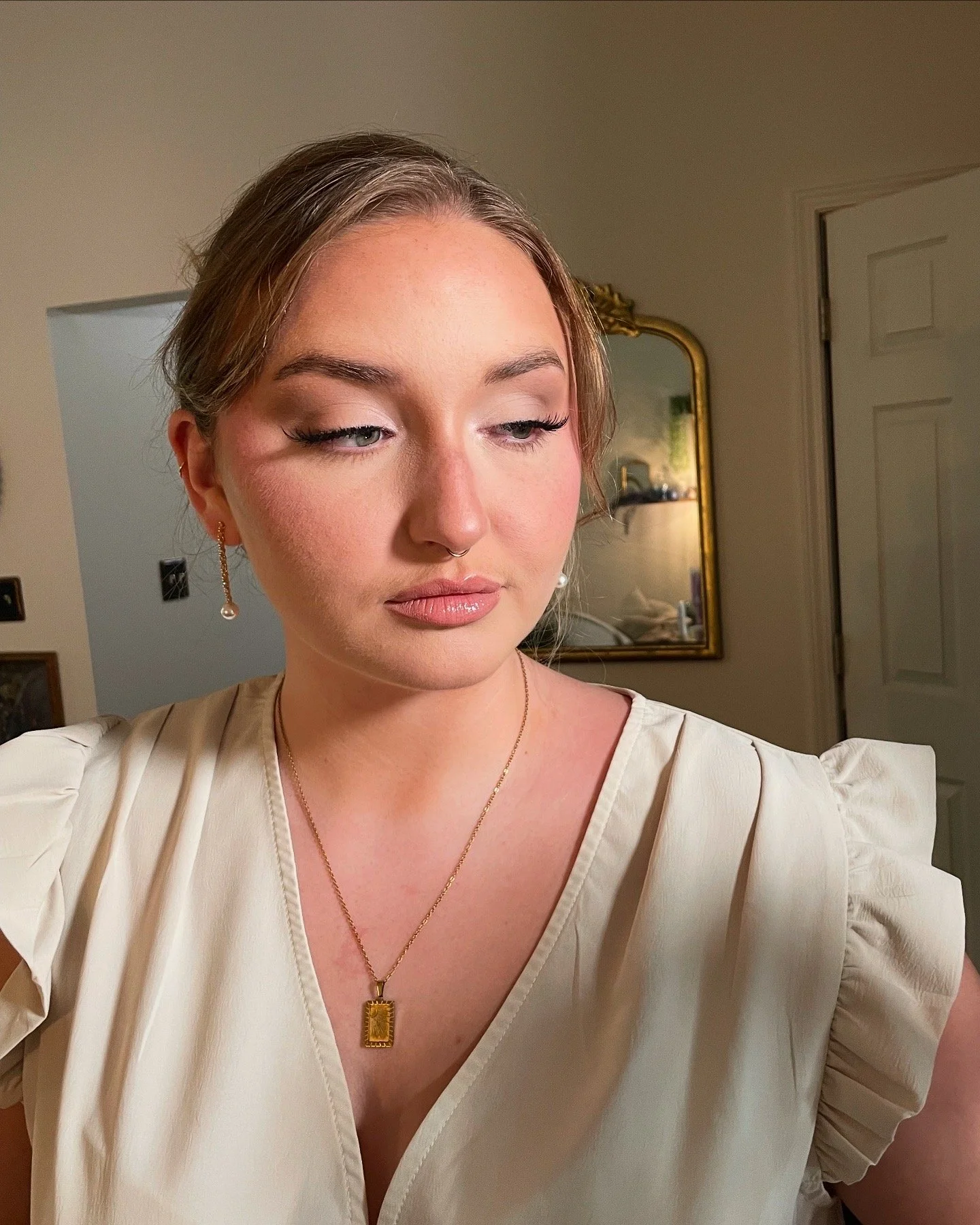A young woman with light makeup and a septum piercing, wearing a cream-colored blouse with puffed sleeves, gold jewelry including dangling pearl earrings and a rectangular pendant necklace, is looking down slightly in a warmly lit room with a mirror 