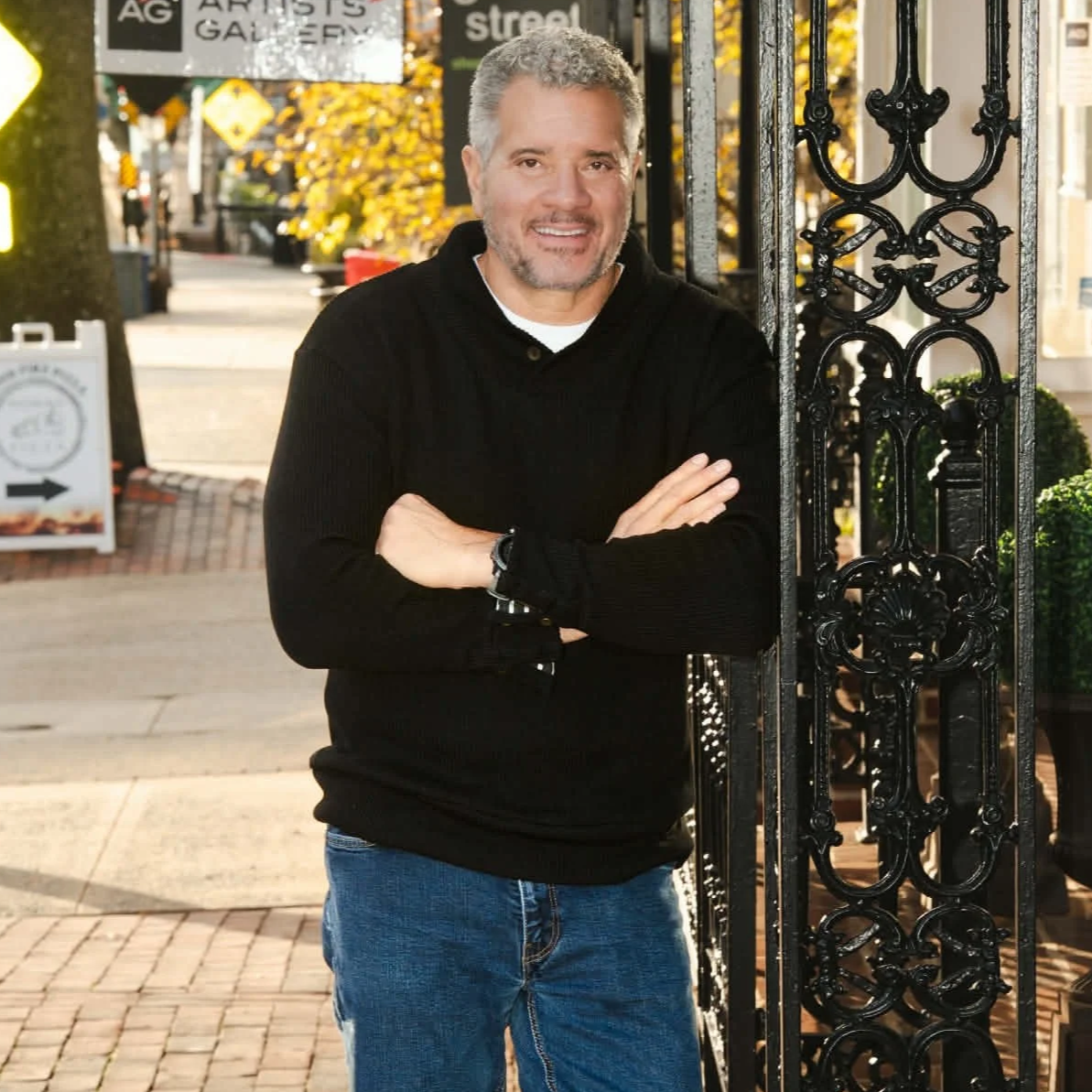 Dwight Woods, Chief Executive, with gray hair and beard, wearing a black hoodie and jeans, standing outdoors next to a decorative black wrought iron gate with arms crossed, smiling at the camera during daytime.