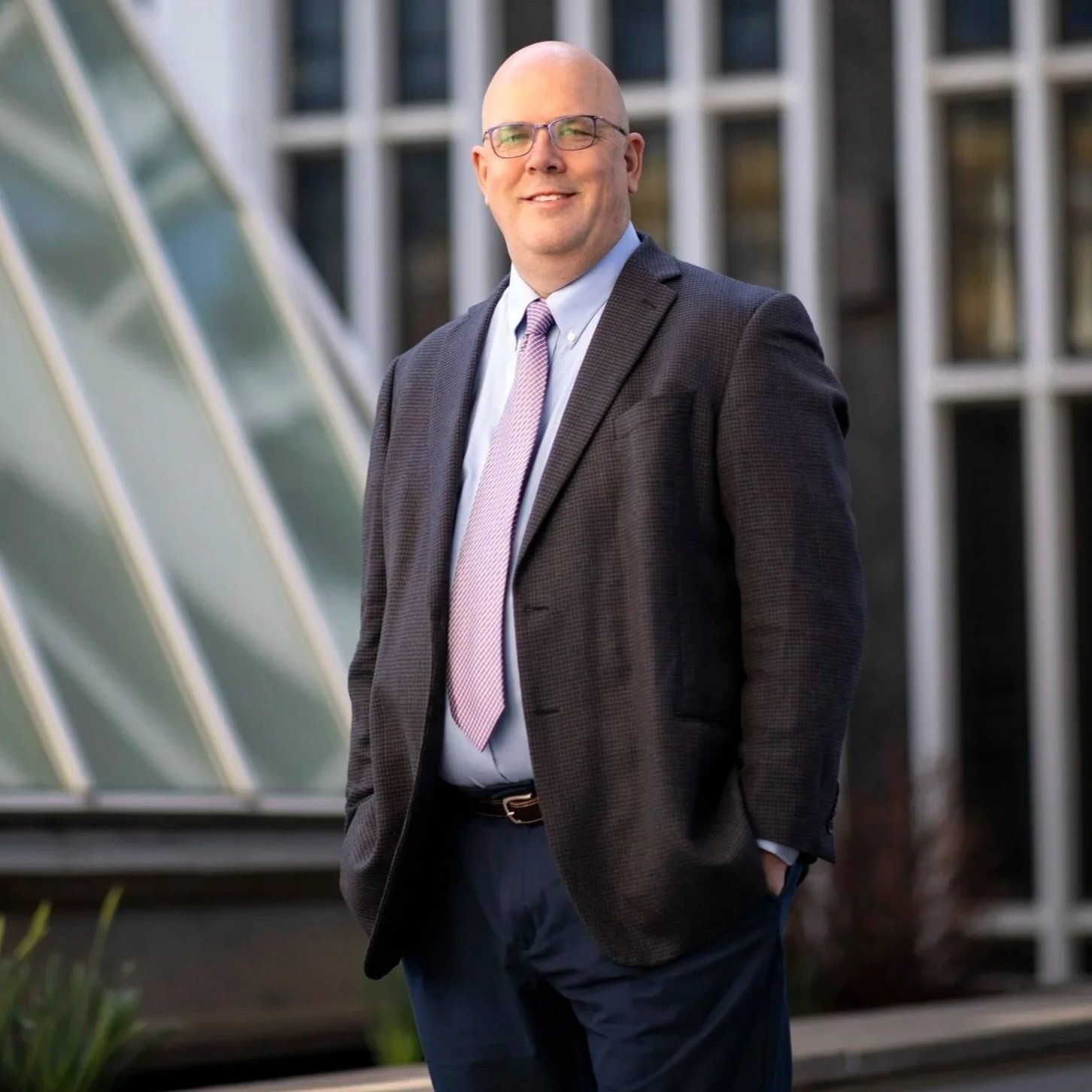 Dr. Shawn Bushway, Chief Scientist, with glasses, wearing a business suit and tie, standing outdoors in front of a modern building with large glass windows.