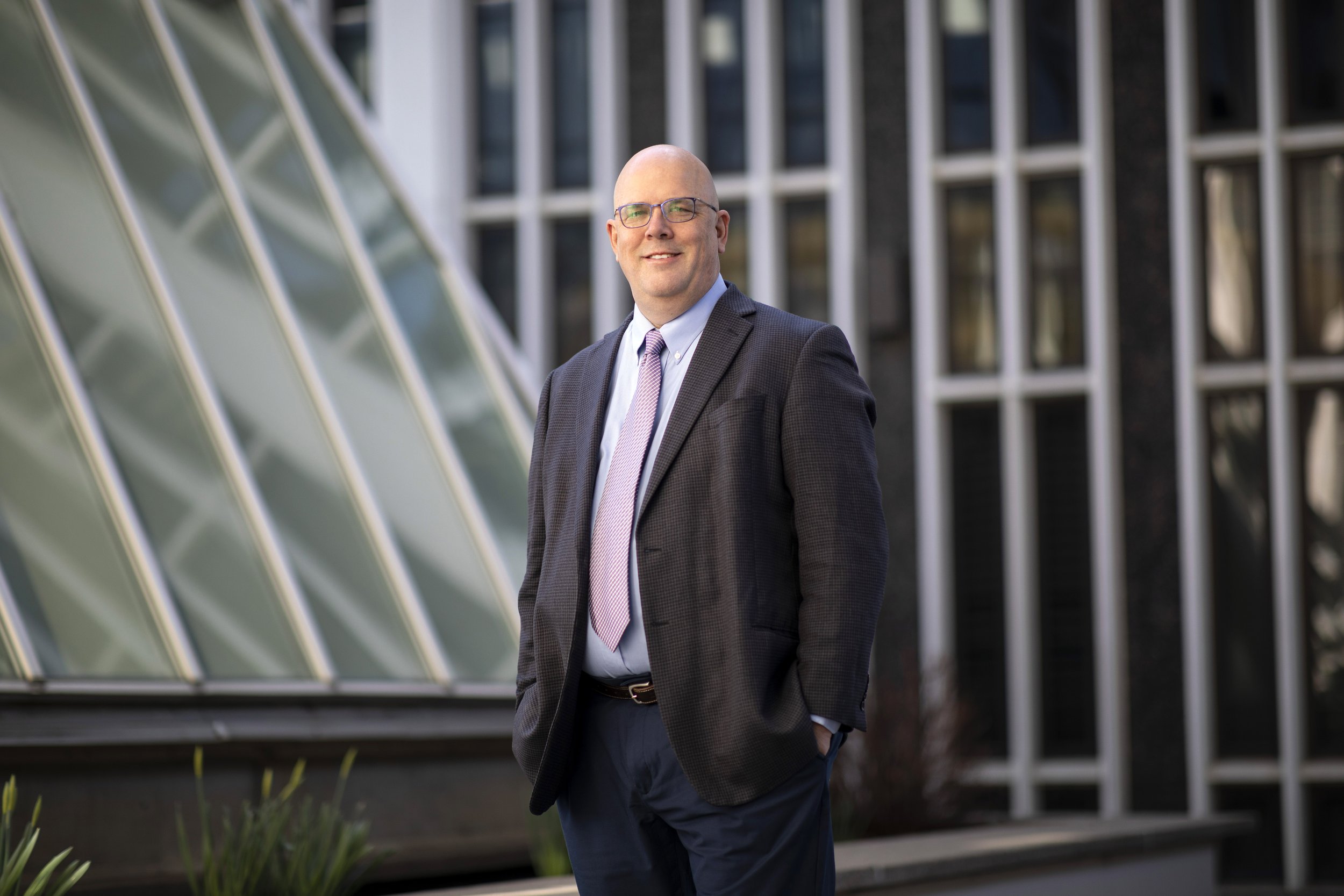 Dr. Shawn Bushway, Chief Scientist, wearing glasses, a dark blazer, white shirt, and pink tie, standing outdoors near a modern building with glass and window panels.
