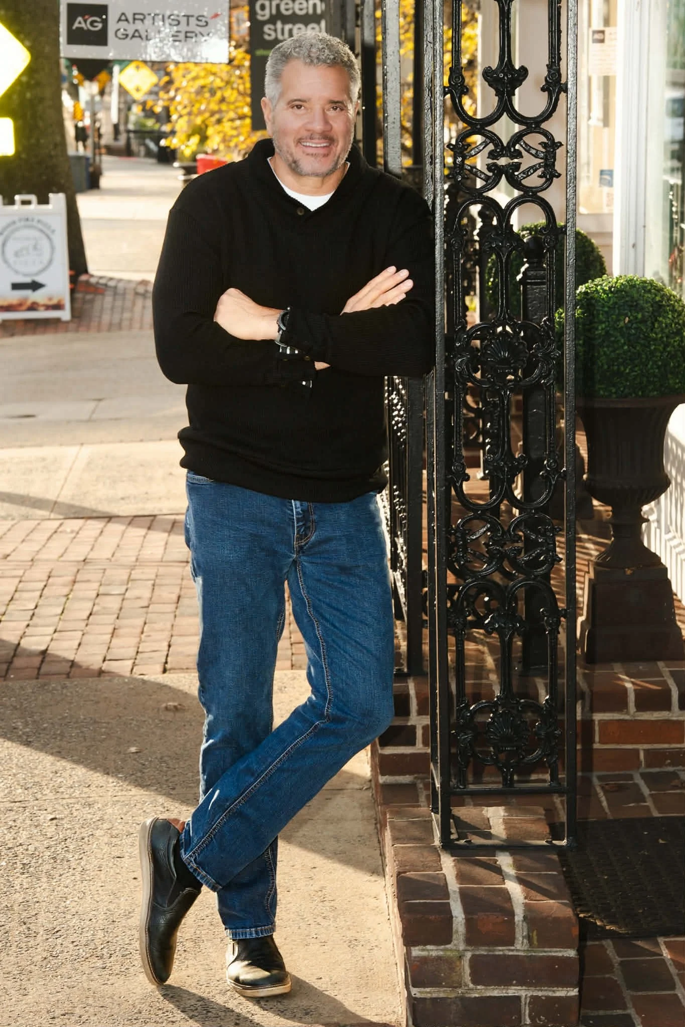 Dwight Woods, Chief Executive, standing next to a decorative black metal gate outside a storefront with a sign that says 'Artists Gallery'. He is smiling, crossing his arms, and leaning slightly against the gate on a brick sidewalk.