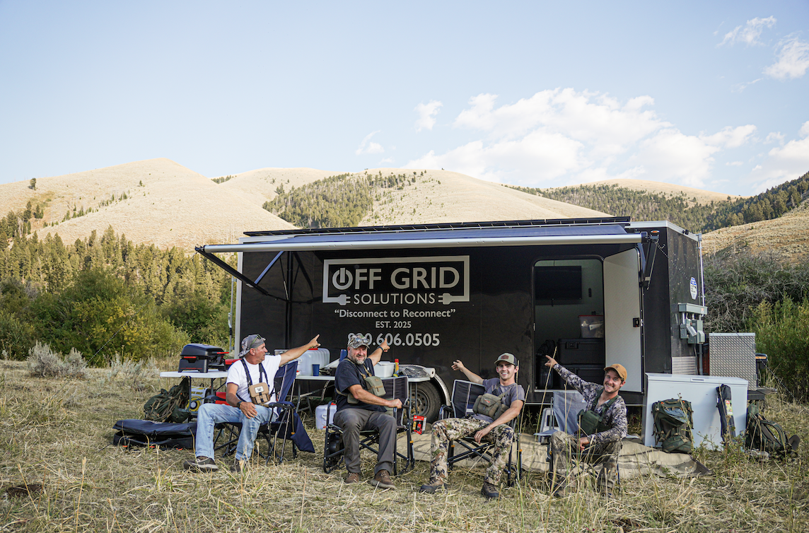 Group of four men in outdoors setting, sitting in camping chairs in front of a mobile office truck labeled "Off Grid Solutions," surrounded by mountains and trees.