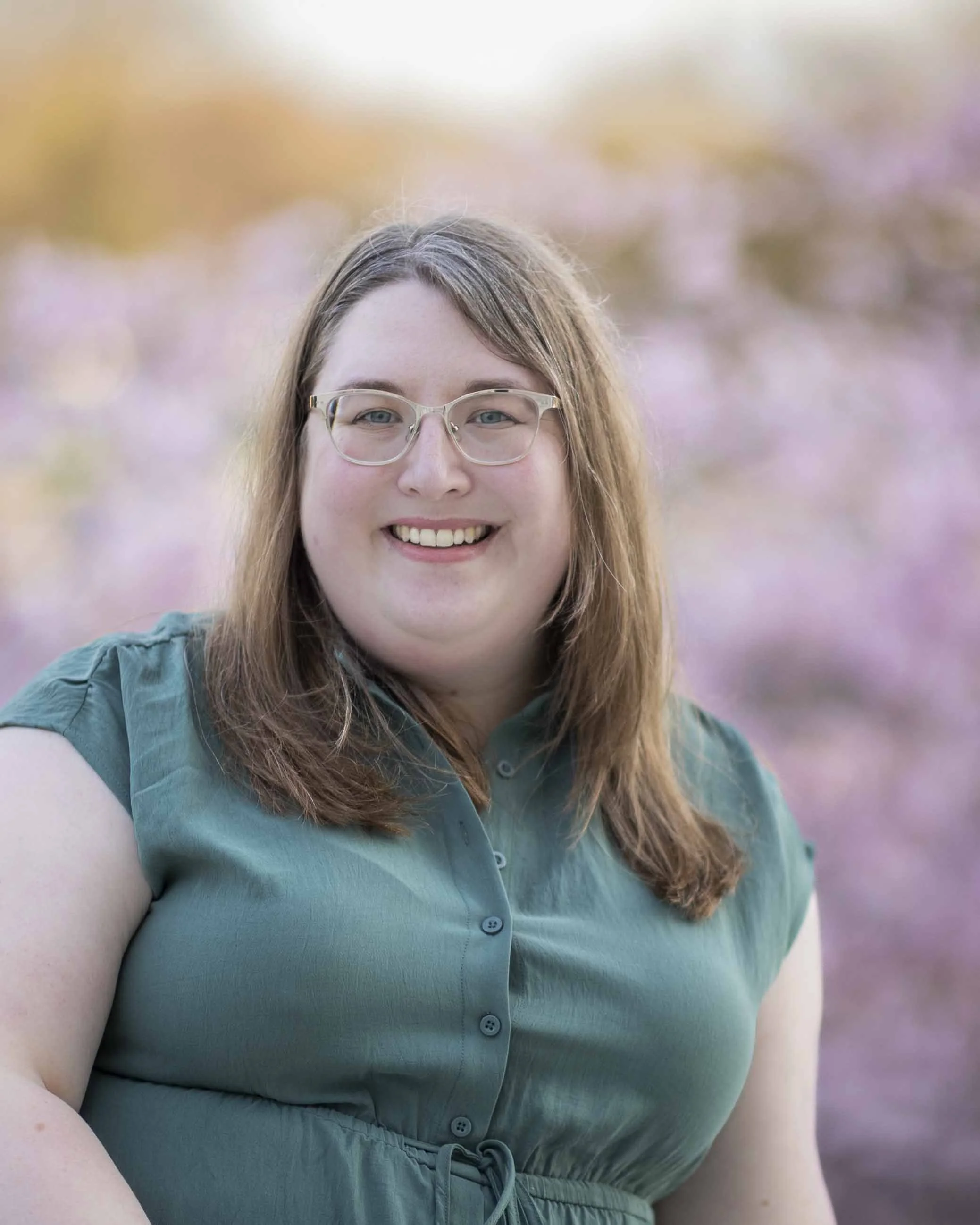A smiling woman with glasses and brown hair standing outdoors in front of a background with pink and purple flowers.
