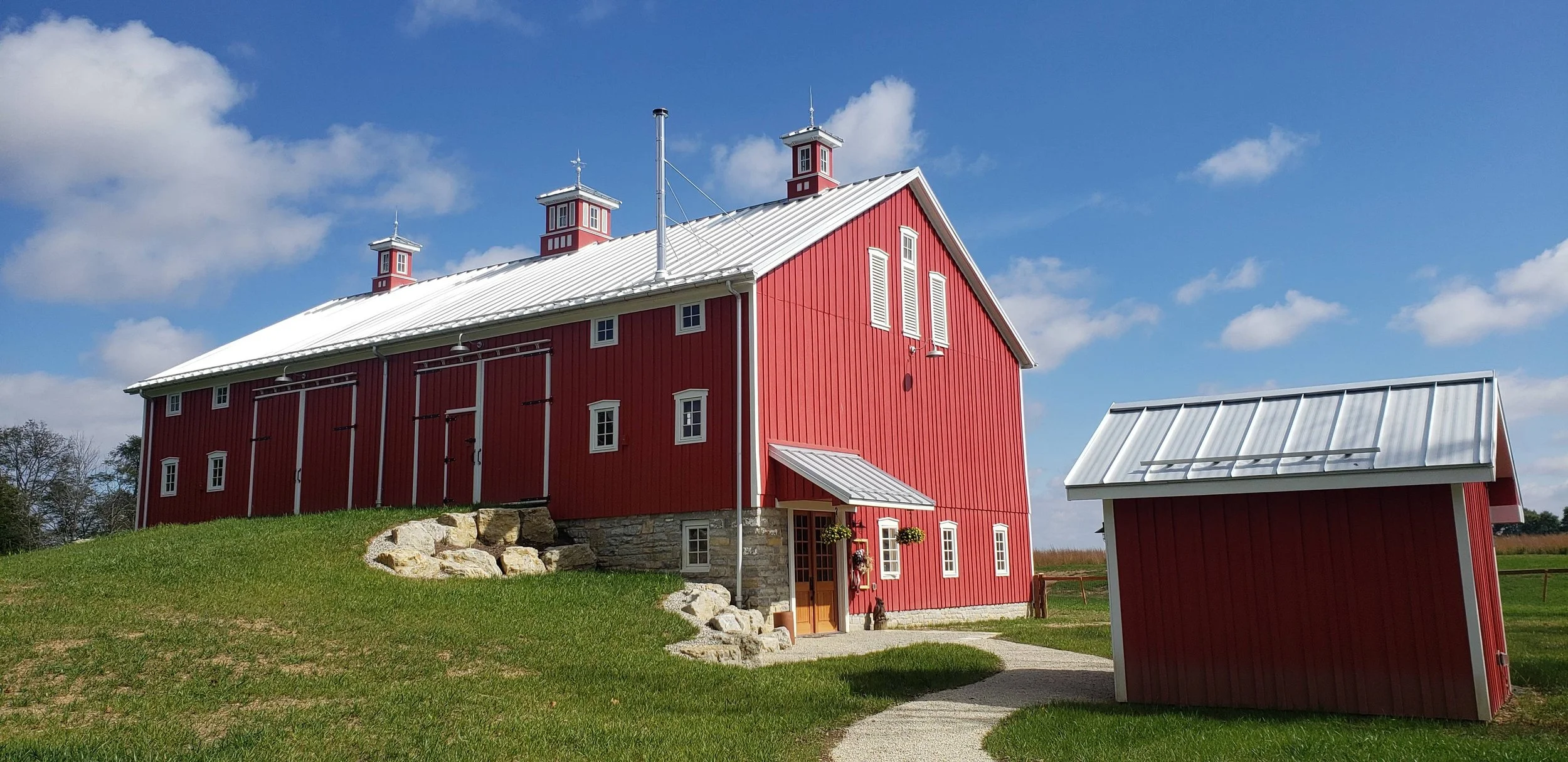 Red barn with white roof on green grassy hillside under blue sky with clouds.