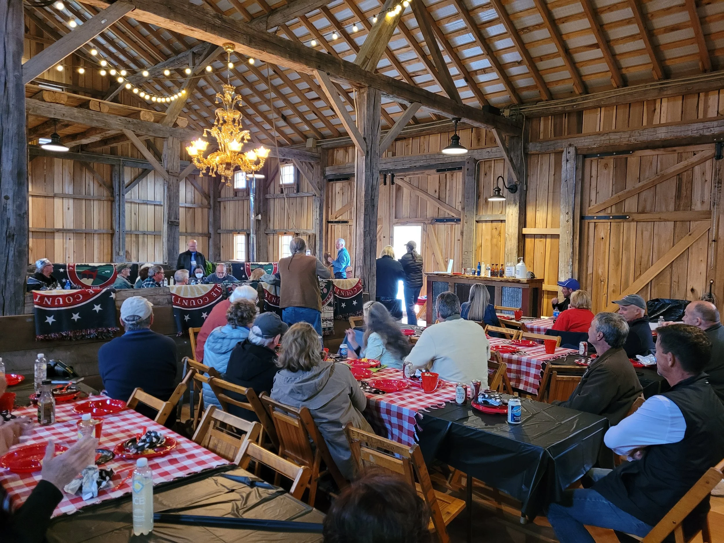 A rustic barn interior with wooden walls, high ceiling, and string lights. People are gathered around tables with red checkered tablecloths, and a chandelier hangs from the ceiling. Some individuals are standing and talking, while others are seated and eating.