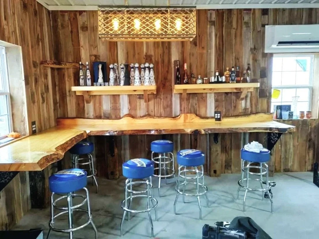 Wooden bar area with five blue stools, bottle display on two shelves, and a window with a view outside.