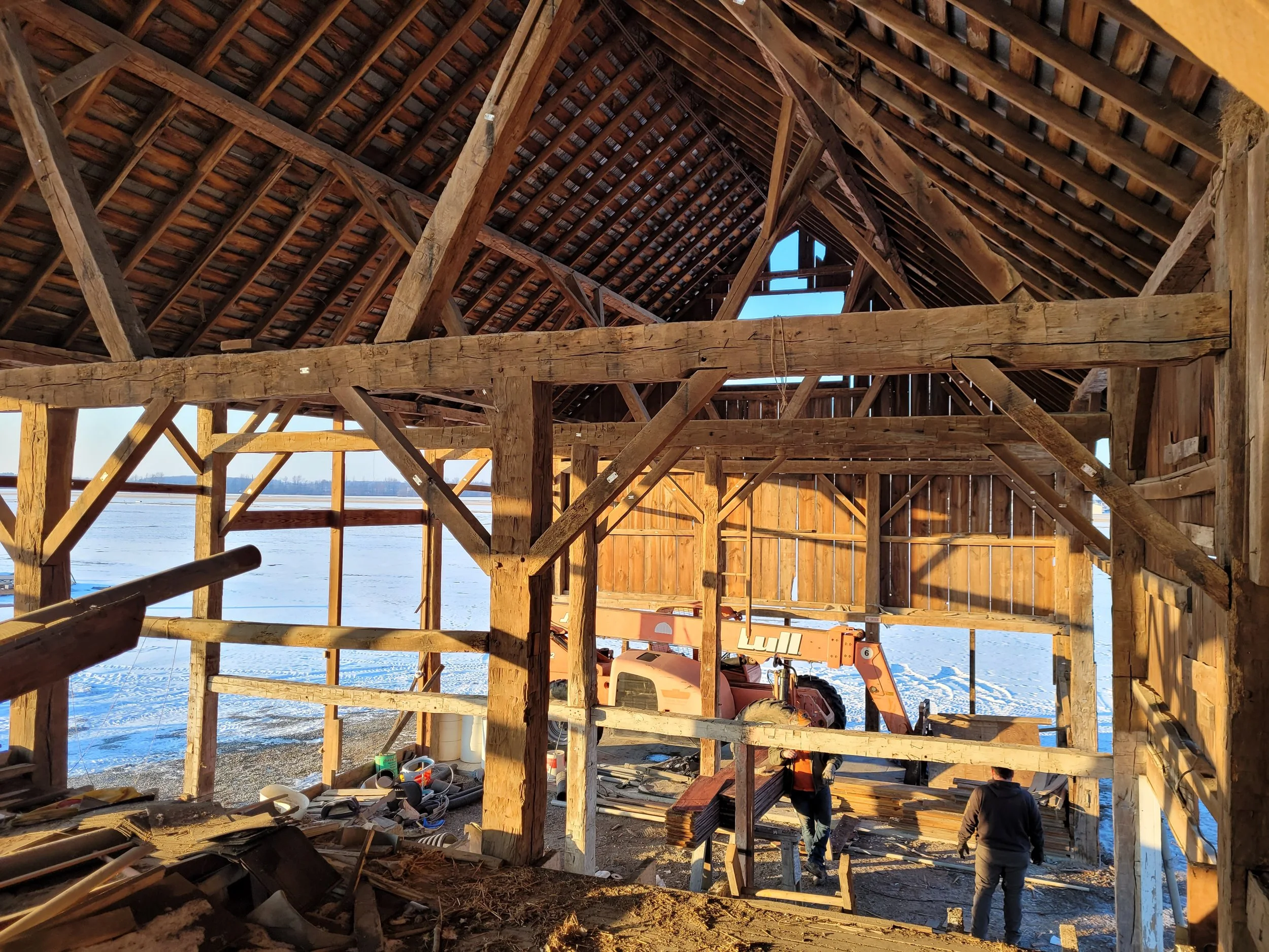 Construction workers building a wooden structure inside a partially built barn with a snowy outdoor landscape visible through the open walls.