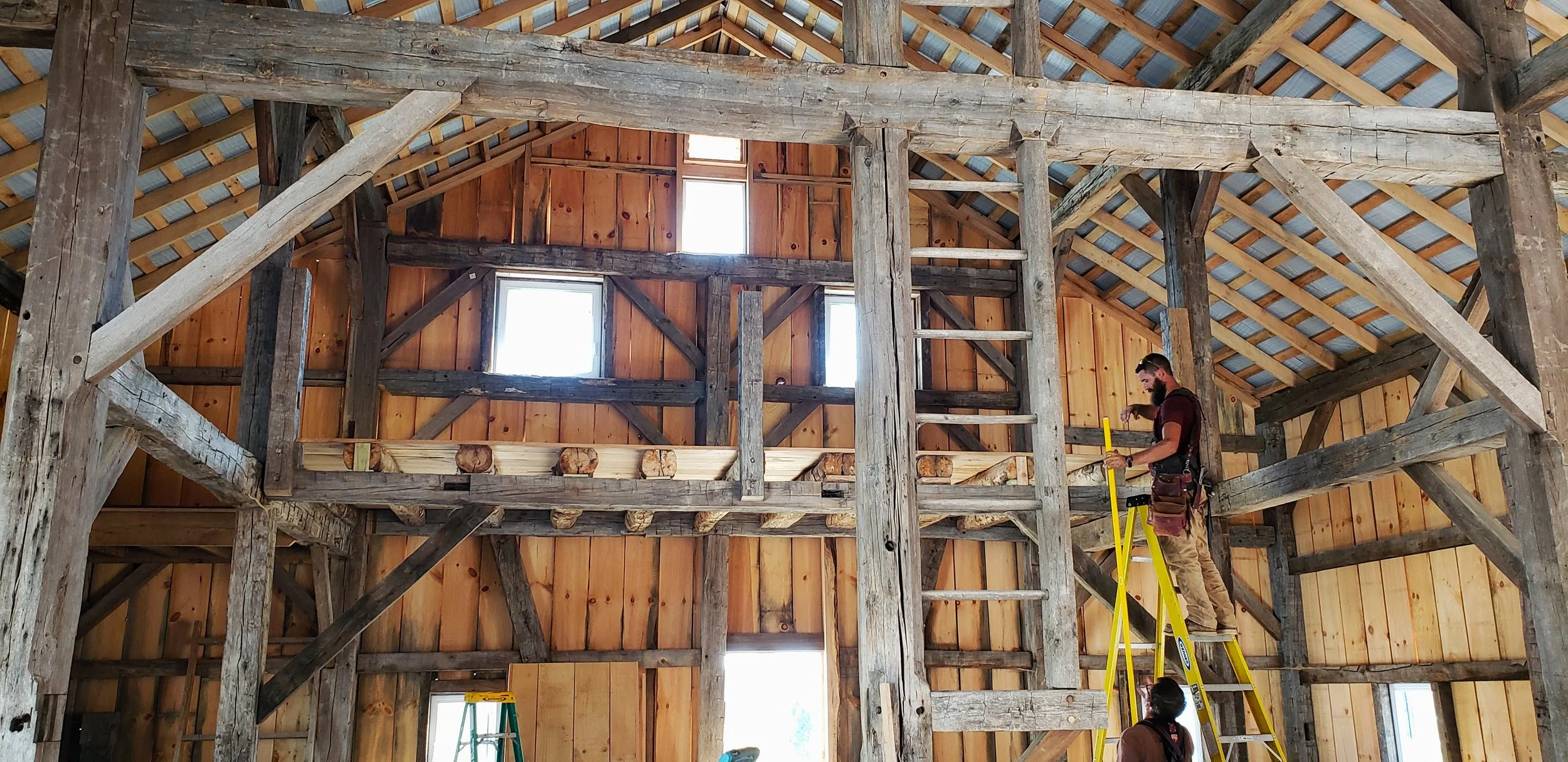 A man standing on a yellow ladder, working on the wooden frame of a barn under construction, with another person at the base assisting.