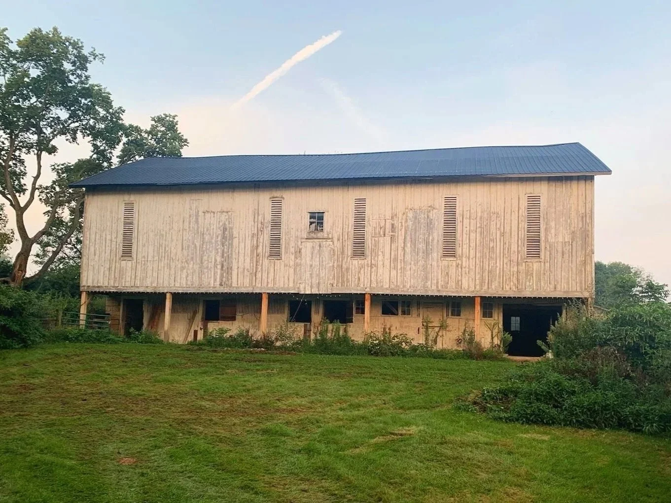 An old wooden barn on stilts with a metal roof surrounded by green grass and trees.