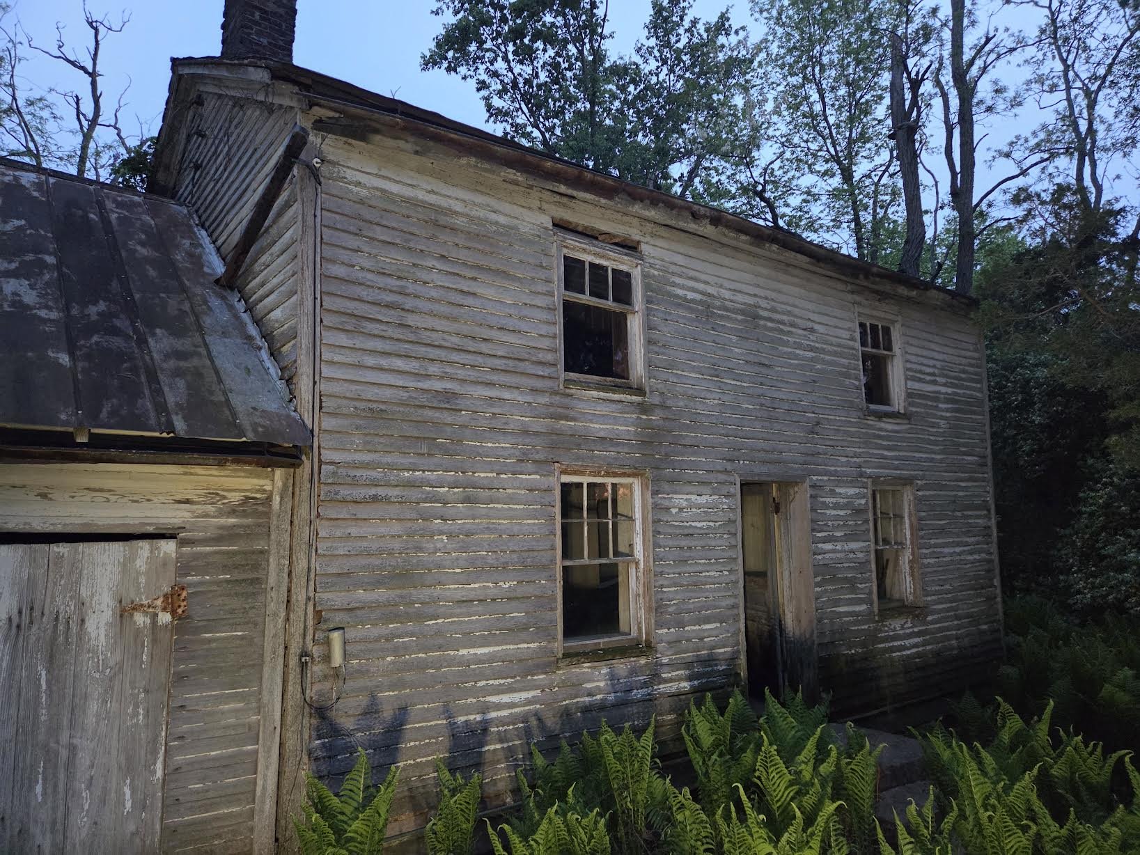 An old, weathered, two-story wooden house with peeling paint and broken windows, surrounded by lush greenery.