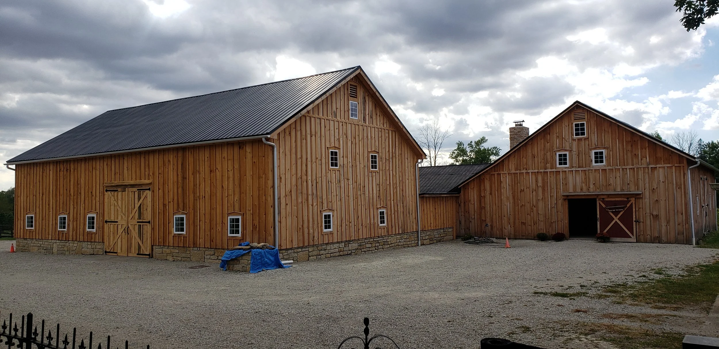 Two large wooden barns with metal roofs under cloudy sky, gravel driveway in front, small windows, and a stone foundation; outdoor equipment and cones nearby.