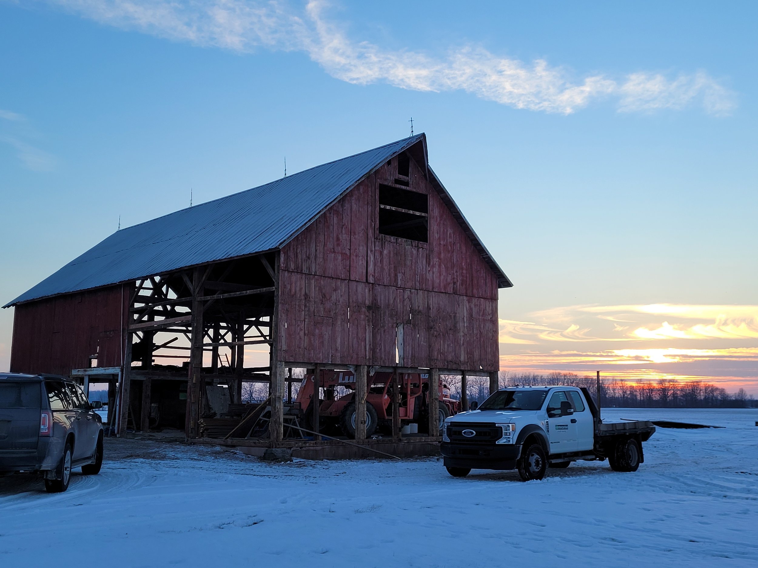 A weathered red barn with a steep metal roof and open upper window stands on a snowy landscape. Several vehicles, including a white pickup truck, are parked nearby. The sky is partly cloudy with the sun setting or rising, casting a warm glow in the horizon.
