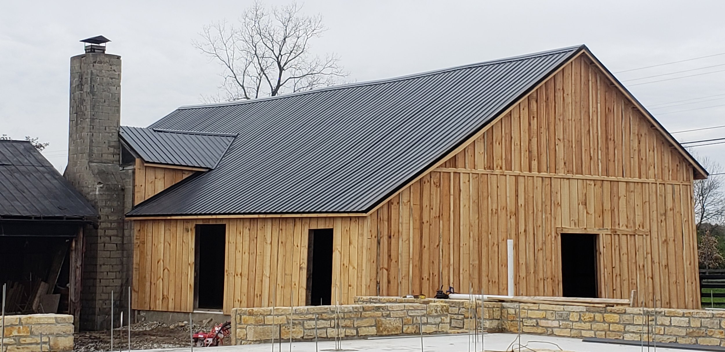 Newly built wooden house with black metal roof, brick chimney, and opening for windows, construction materials around, overcast sky.
