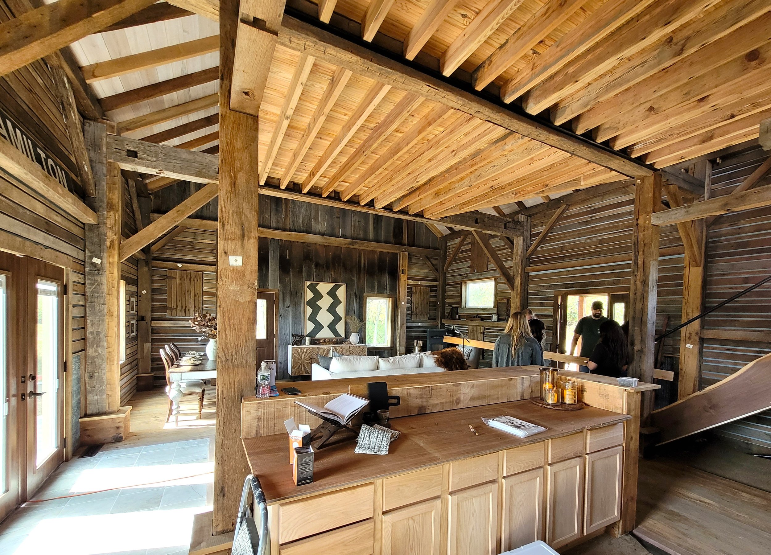 Open interior of a rustic wooden home or barn with people gathered near a staircase, wooden beams, and a dining area with chairs and a table.