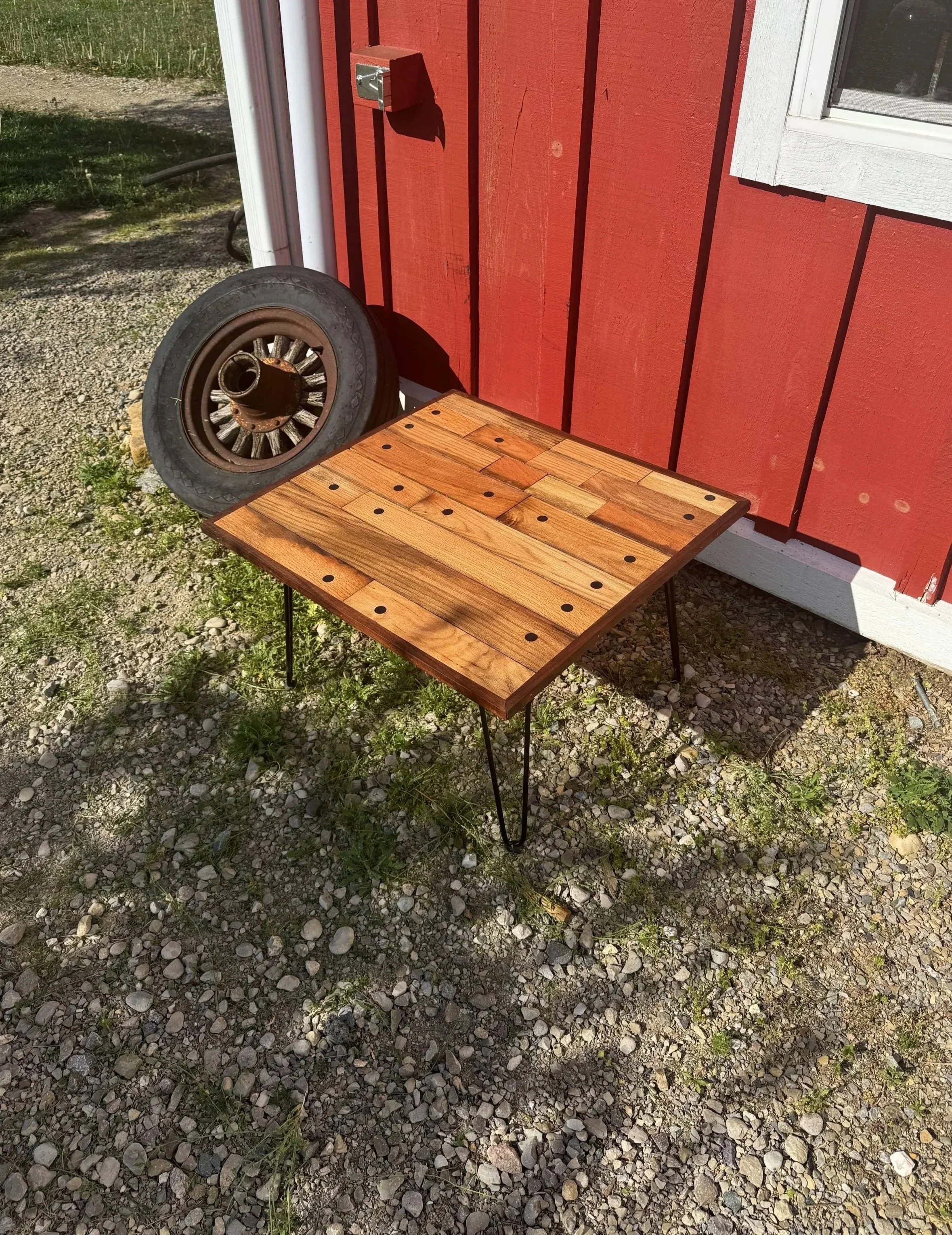 A foldable wooden table with black metal legs outside on a gravel surface near a red wooden building. An old car wheel with a rusted hubcap is leaning against the building.