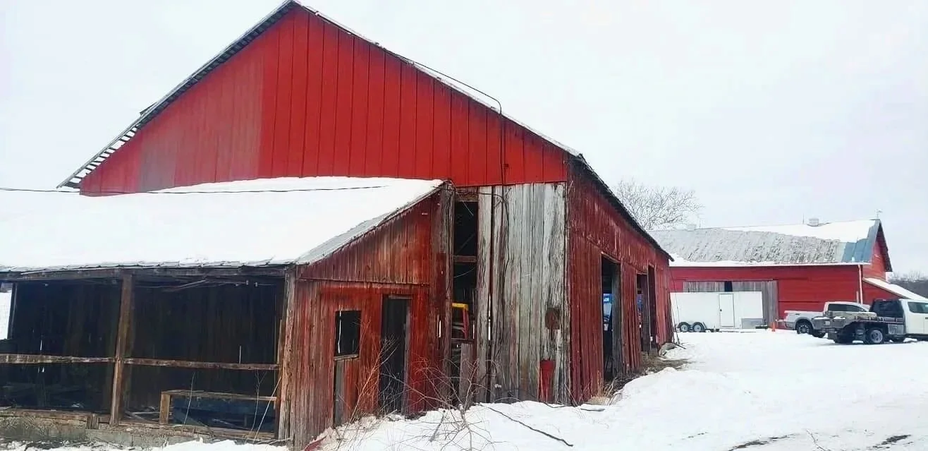 Red and weathered barn and shed covered in snow with trucks parked nearby in a winter landscape.