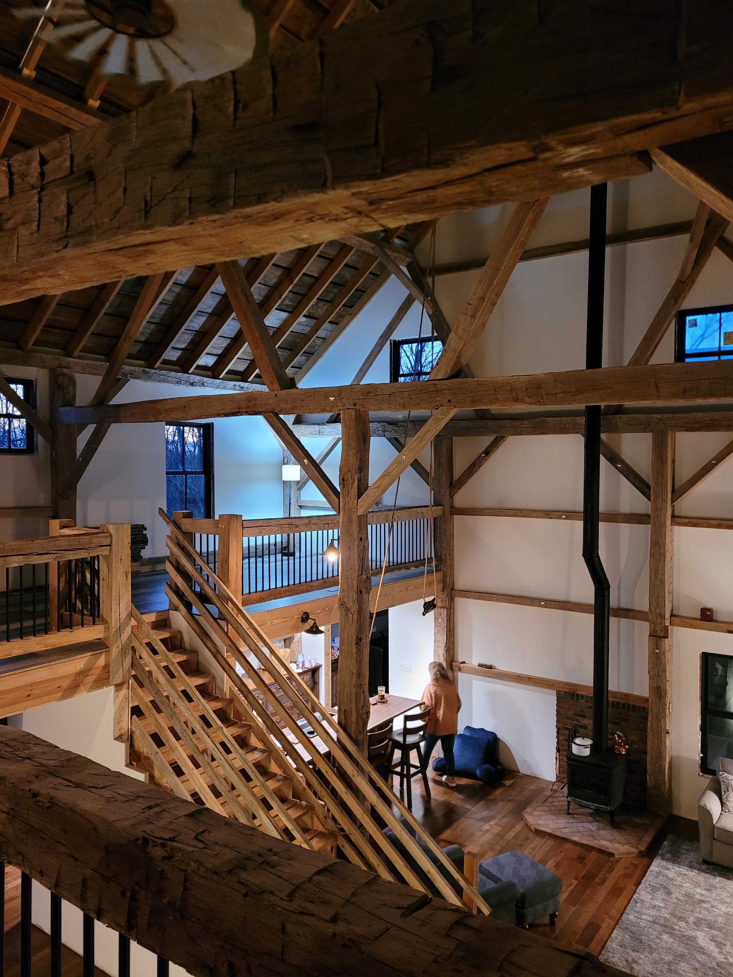 Interior view of a rustic home with exposed wooden beams and a staircase, a person standing at a dining table, a wood stove with a chimney pipe, and windows showing a winter scene outside.