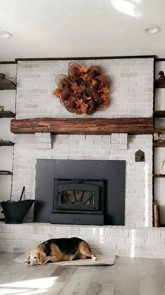 A cozy living room with a white brick fireplace, decorated with fall-themed wreath above the mantel. A dog is sleeping on a small mat in front of the fireplace on the wooden floor.