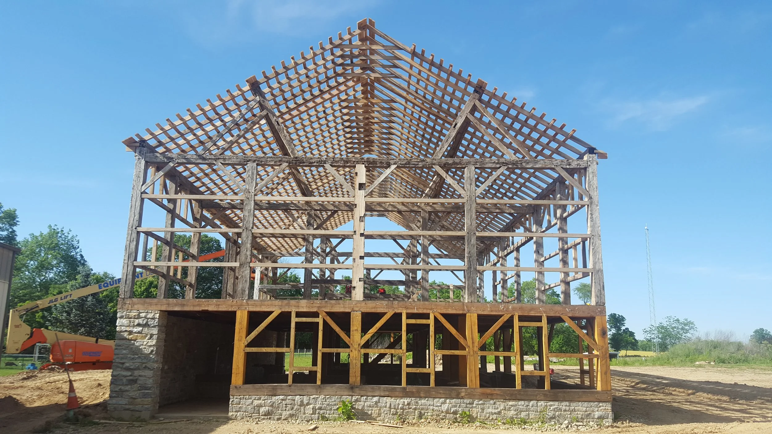 Wooden house under construction with roof framing, supported by wooden beams, on a raised brick foundation, with construction equipment nearby and trees in the background.