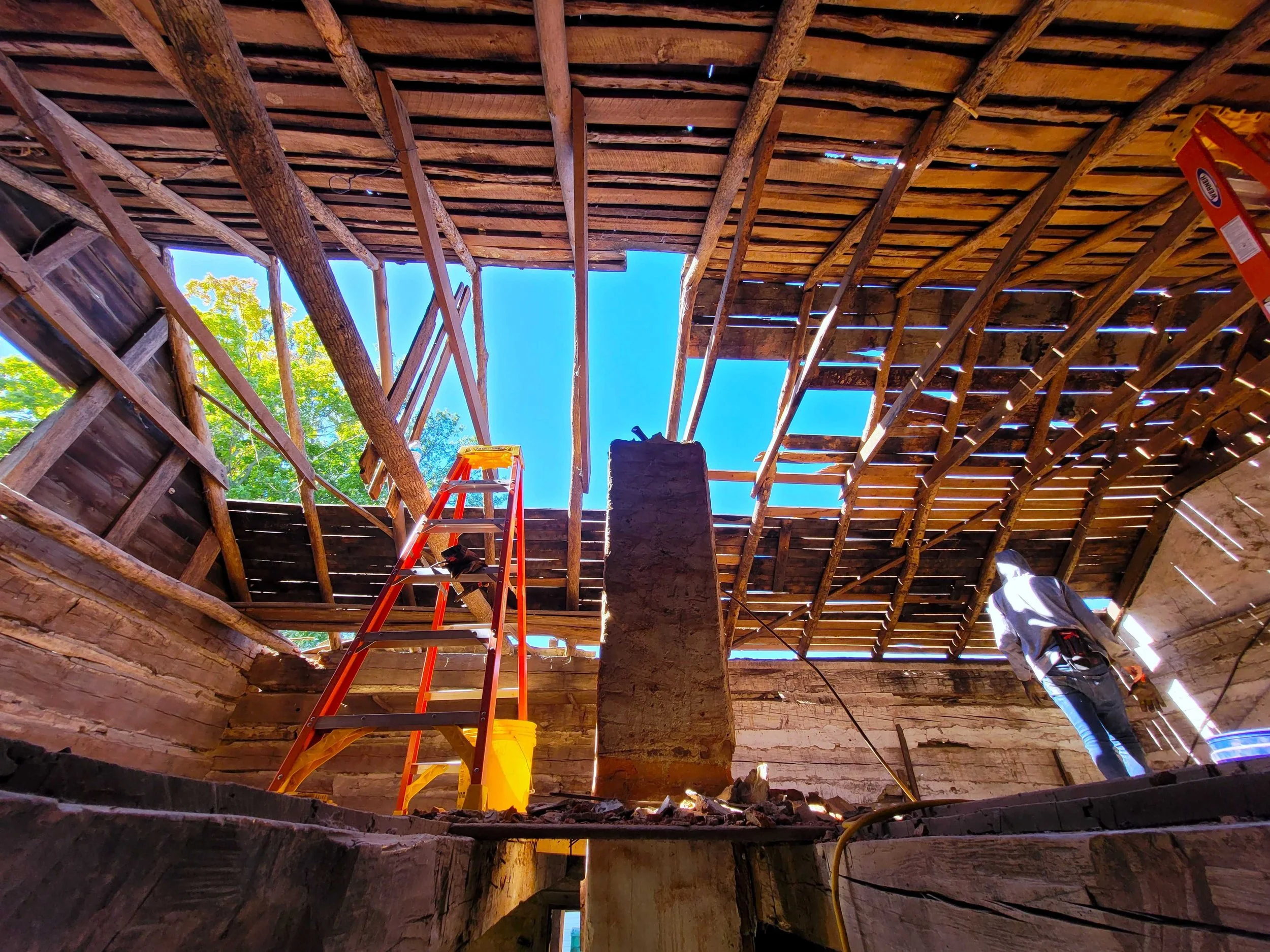 View from inside a building under construction with a ladder, exposed wooden beams, a person working, and an opening in the roof revealing a bright blue sky and trees.