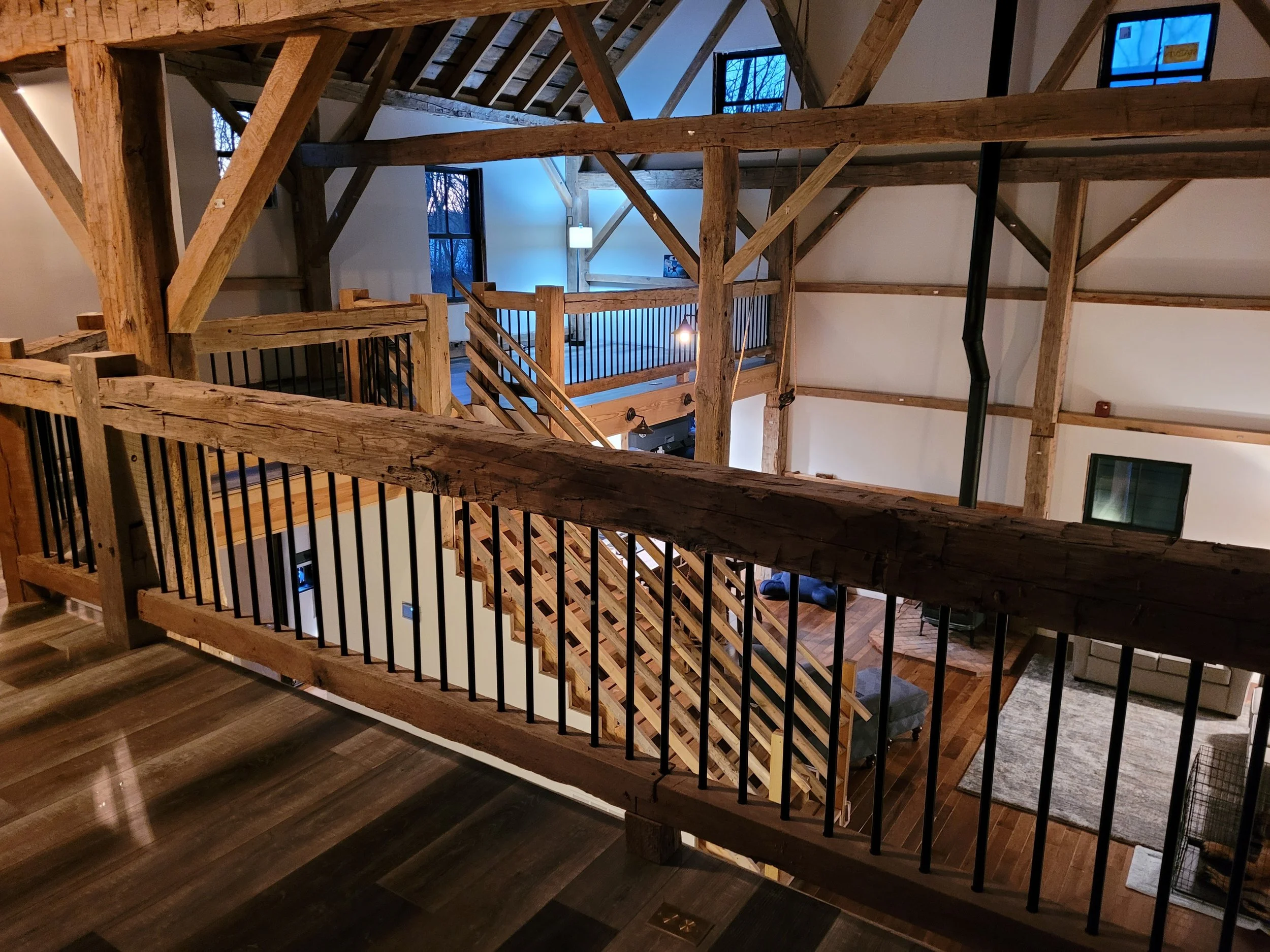 Interior view of a multi-level staircase with rustic wooden beams and black metal railings, illuminated by indoor lighting and seen against dark blue evening sky through the windows.