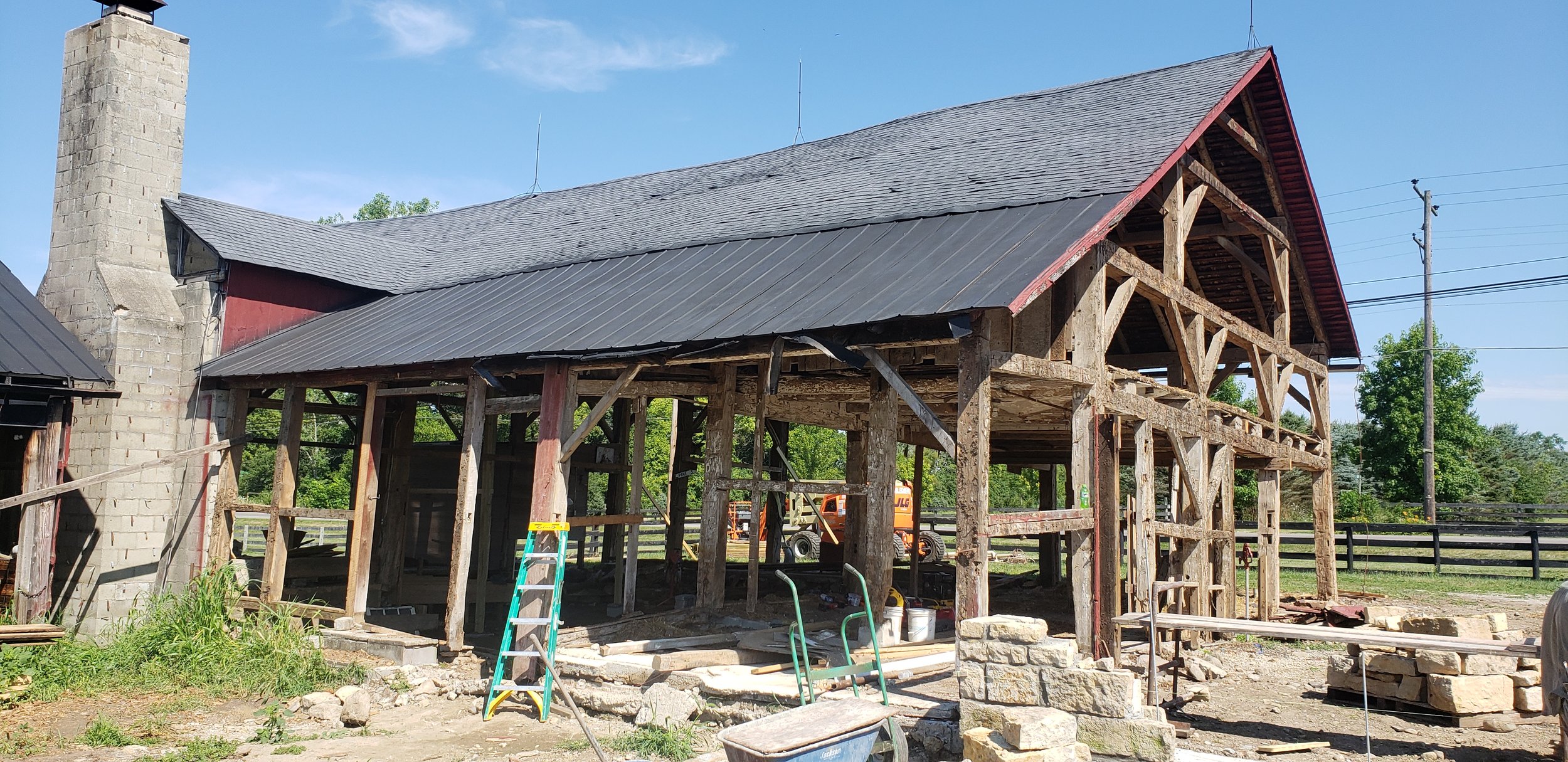 A barn under construction with a partially built wooden structure, a metal roof, and a brick chimney. Construction equipment and materials are visible nearby.