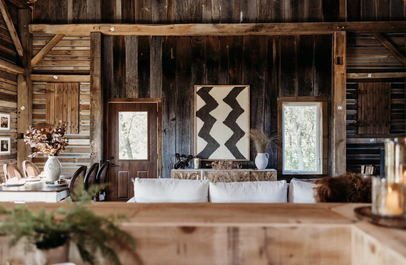 Interior of a rustic wooden cabin living room with a white sofa, abstract artwork, and windows showing outdoor trees.