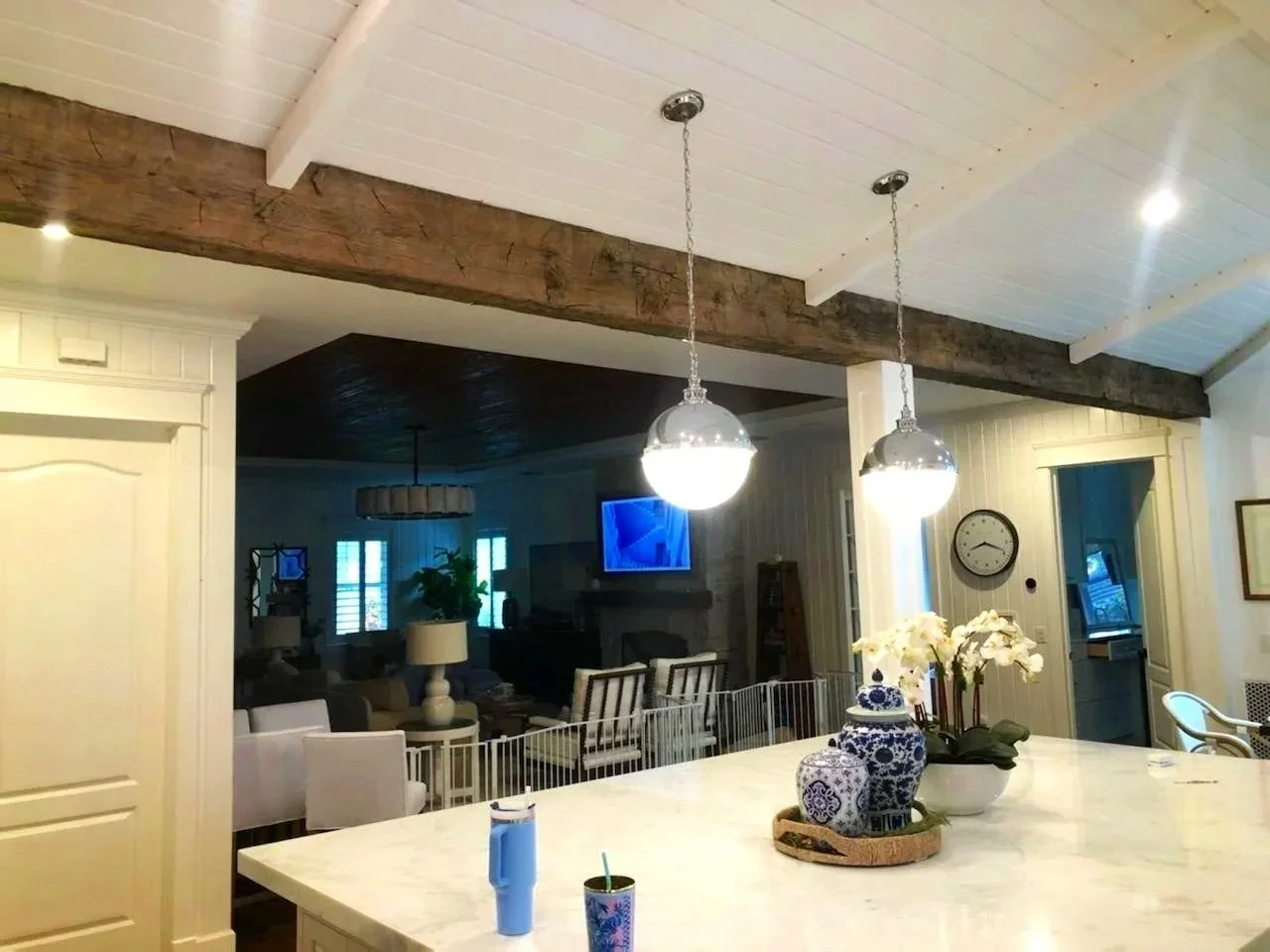 Open-concept living and kitchen area with white cabinetry, a marble countertop, decorative blue and white vases, and hanging pendant lights. The living space features a sofa, chairs, a wall-mounted TV, and large windows.