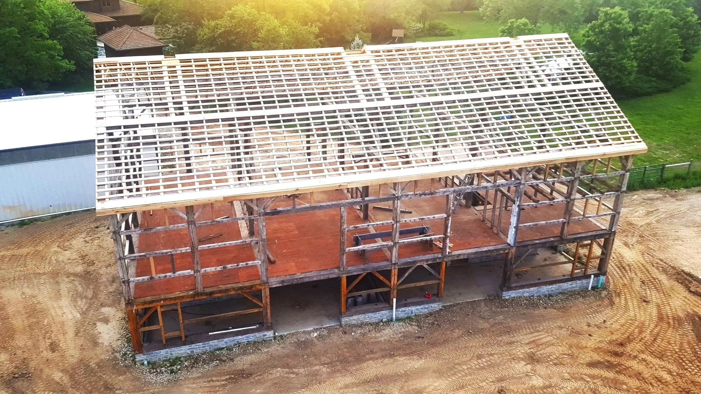 Aerial view of a house under construction with a wooden framework and a partially completed roof, surrounded by dirt and greenery.
