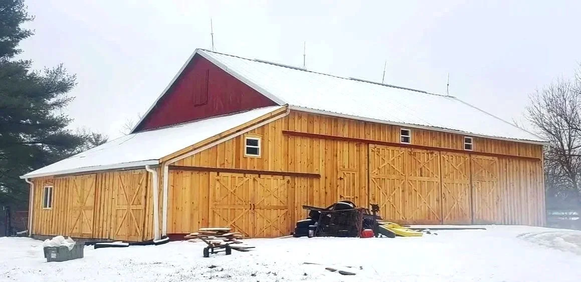 A large wooden barn in a snowy landscape, with a red upper section and four sliding doors, some small windows, and outdoor equipment in front.