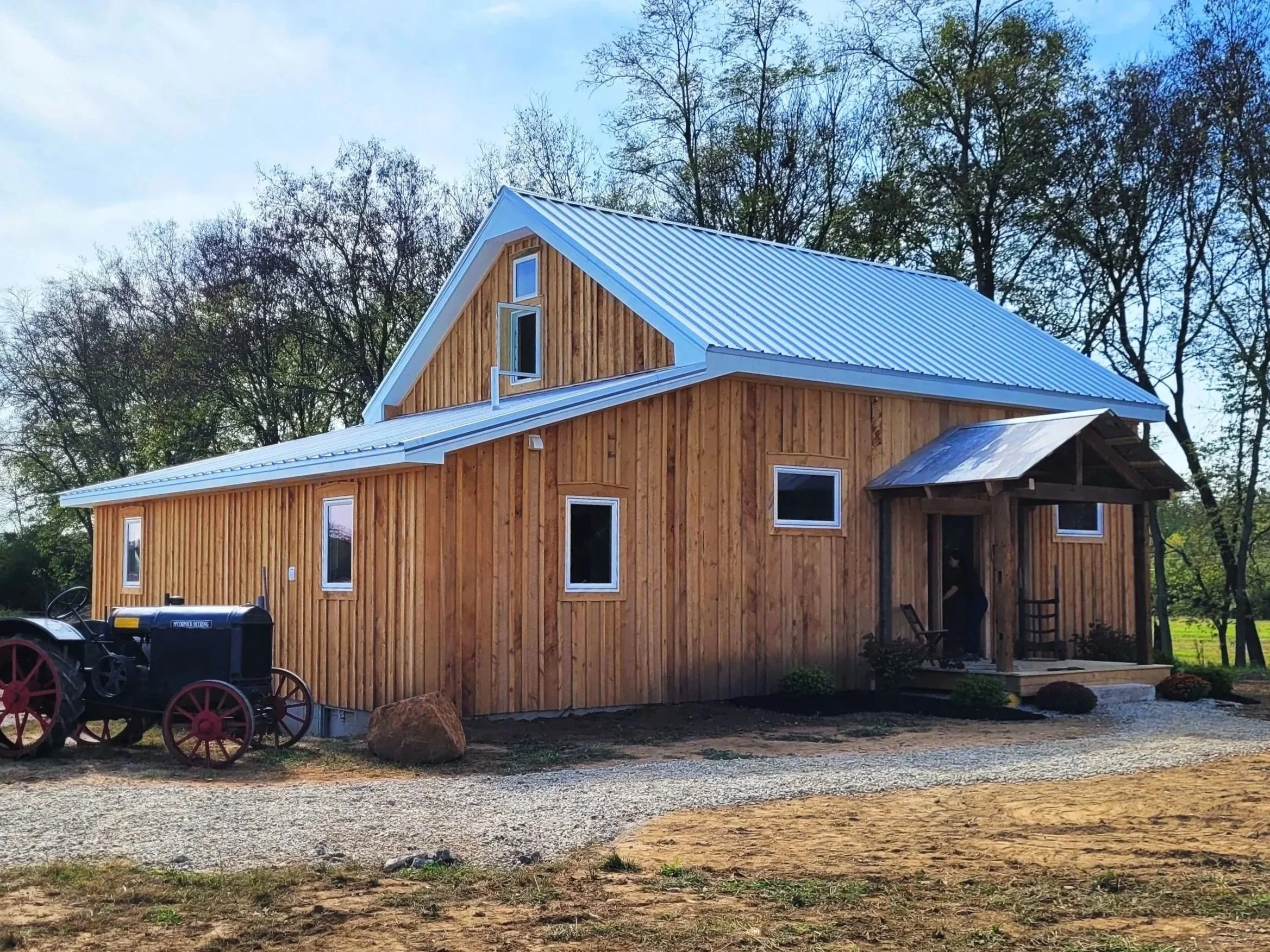 A newly built wooden house with a metal roof, situated on a gravel driveway, surrounded by trees.